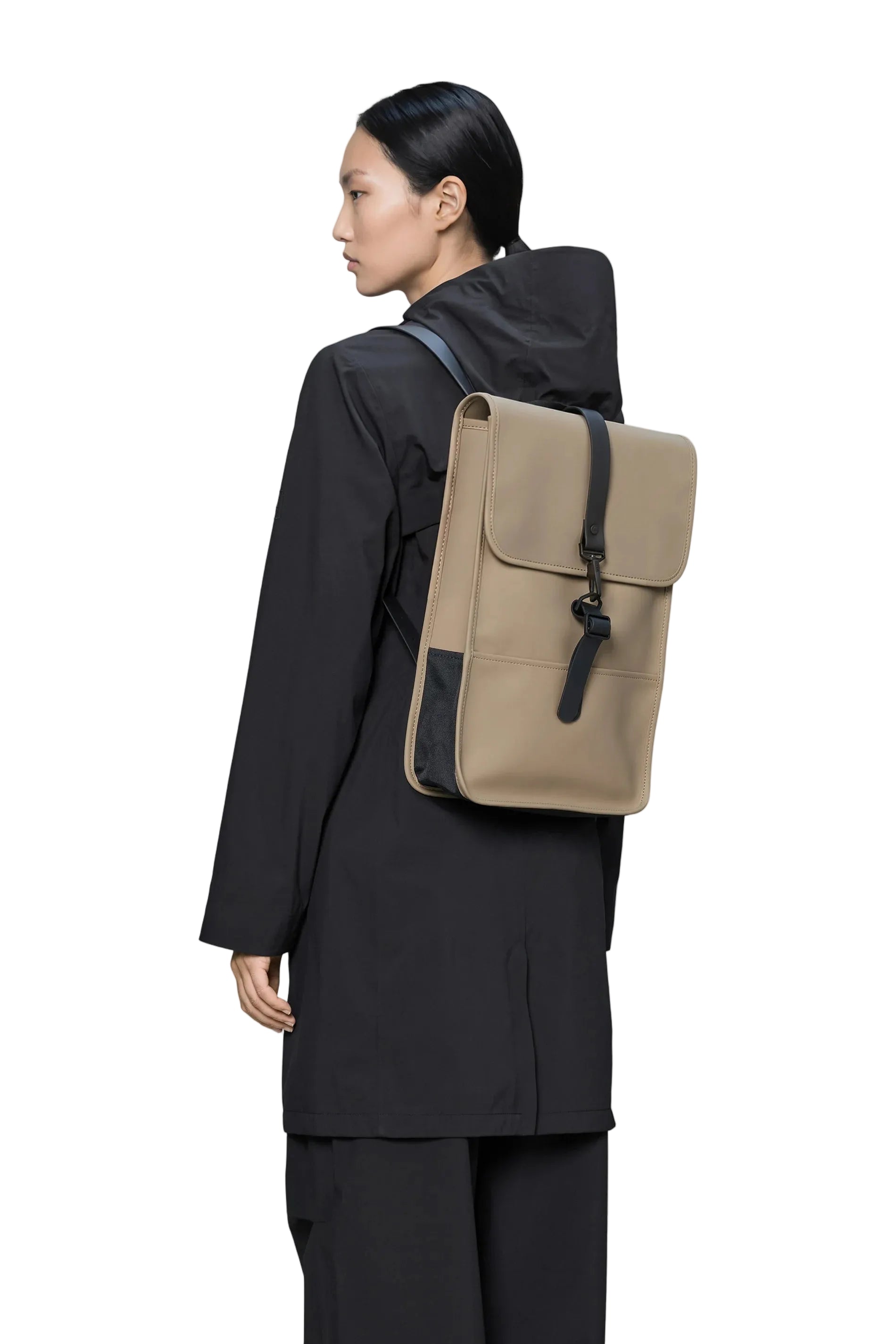 A person, facing sideways and dressed in a black coat and black pants, is carrying the Rains Backpack Mini Bag in Beige by Rains—a rectangular commuter rucksack with a black strap and buckle—against a white background.