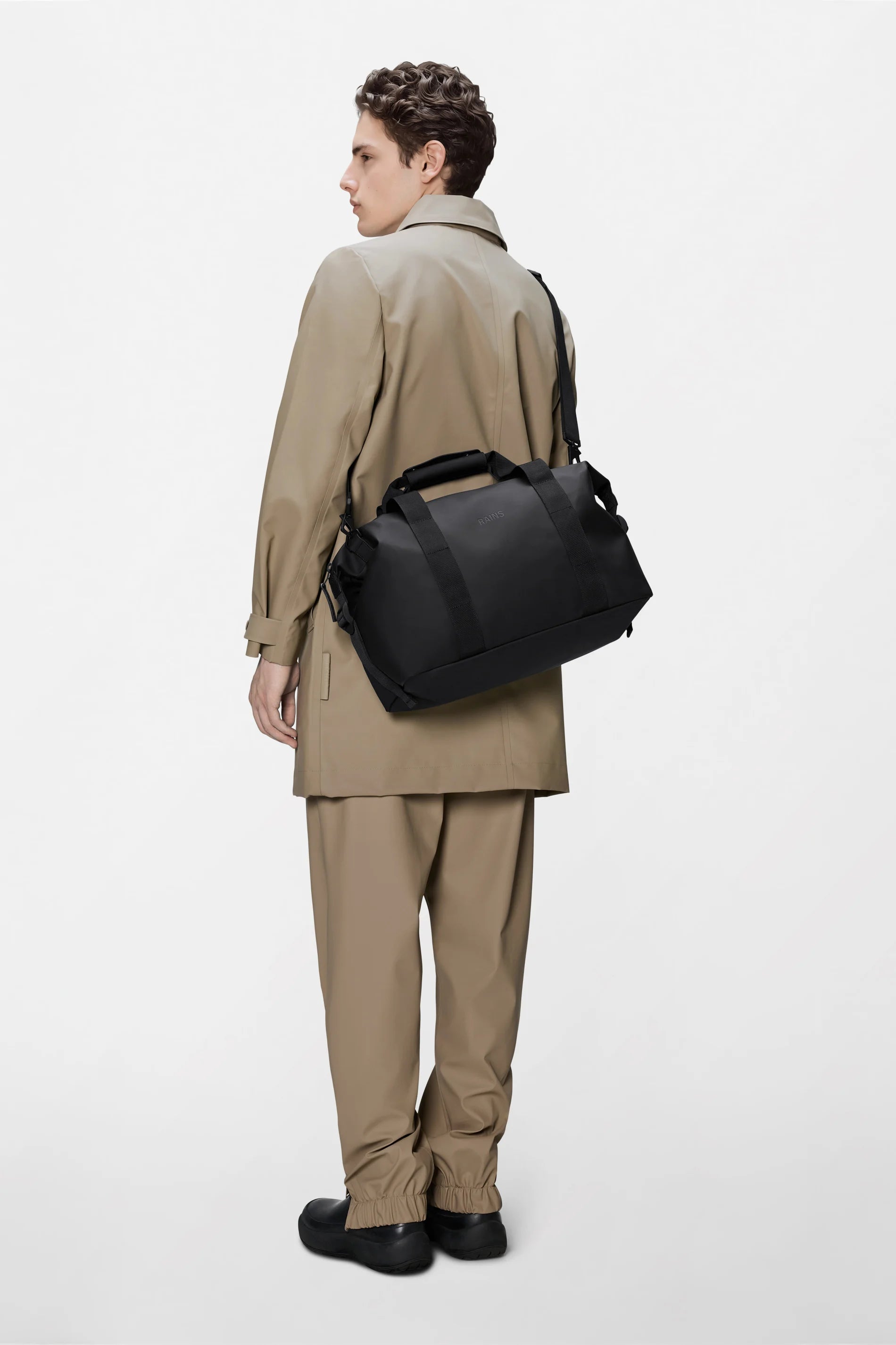 A person wearing a tan trench coat and matching pants stands facing away, carrying the Rains Hilo Weekend Bag Small in Black by Rains over their shoulder against a plain white background.