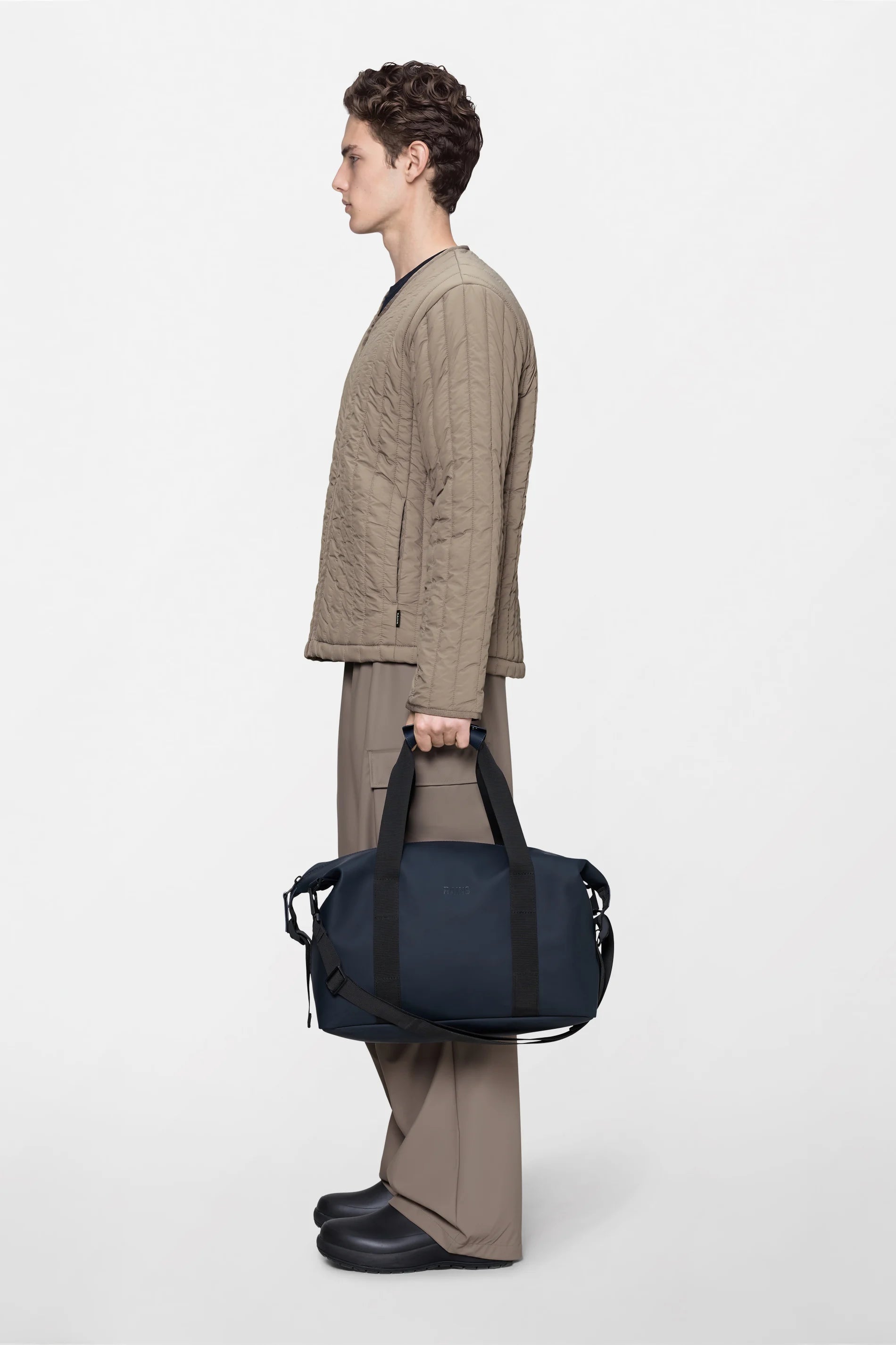 A person in a beige quilted jacket and wide-leg pants stands in profile, holding the Rains Hilo Weekend Bag Small in Navy by the handles against a plain light background.