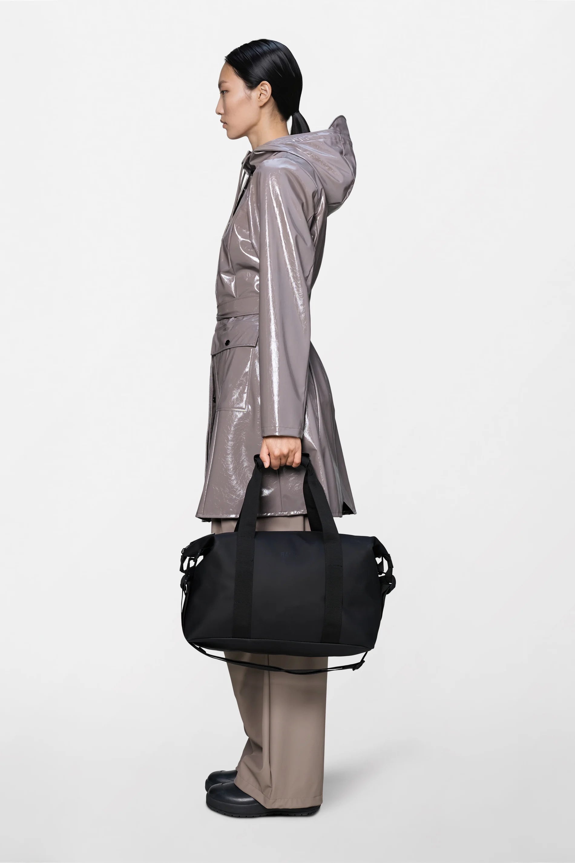 A person stands in profile against a plain white background, wearing a shiny taupe raincoat with a hood, matching taupe pants, and black shoes, while holding the Rains Hilo Weekend Bag Small in Black by Rains.