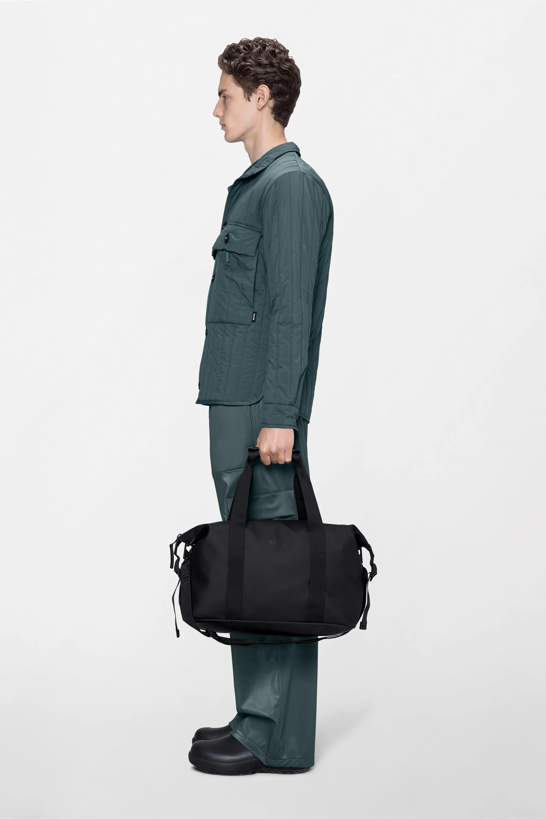 A person in a green jacket and pants stands in profile, holding the Rains Hilo Weekend Bag Small in Black by the handles. The background is plain and light-colored.