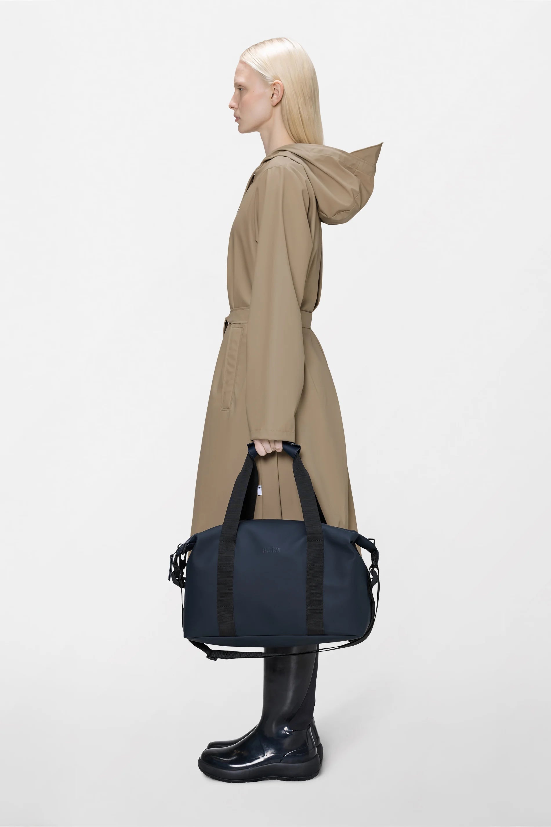 A person in a tan hooded coat and black rain boots stands in profile, holding the Rains Hilo Weekend Bag Small in Navy against a plain white background.