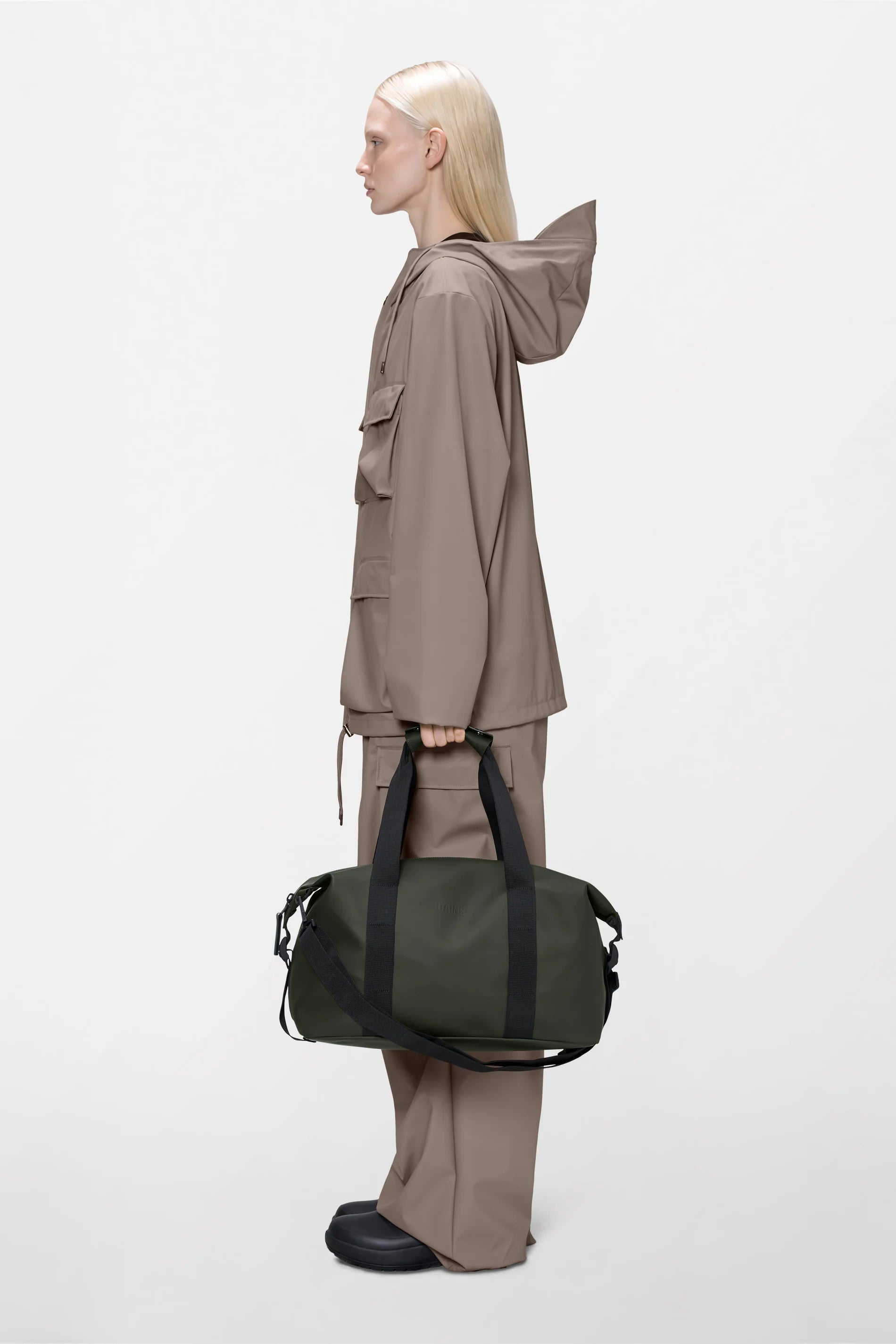 A person with straight blond hair, dressed in a taupe rain jacket and matching pants, stands in profile against a plain white background holding the Rains Hilo Weekend Bag Small in Green—a waterproof overnight bag with black straps from Rains.