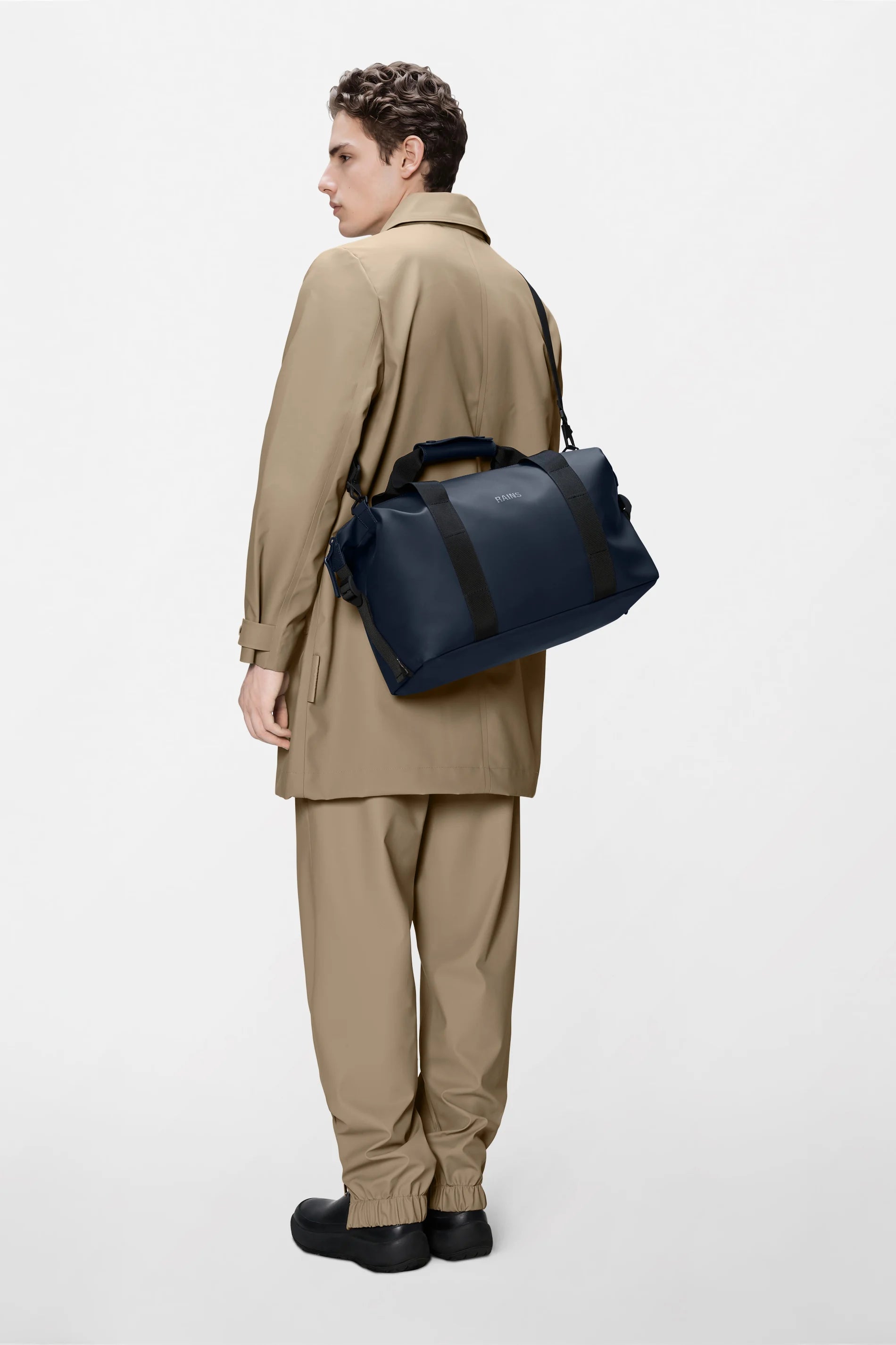 A person stands facing away, wearing a beige trench coat and matching pants with black shoes, carrying the Rains Hilo Weekend Bag Small in Navy over their shoulder. The sleek waterproof bag from Rains, featuring black straps, is set against a plain white background—making it the ideal small travel companion for any quick getaway.