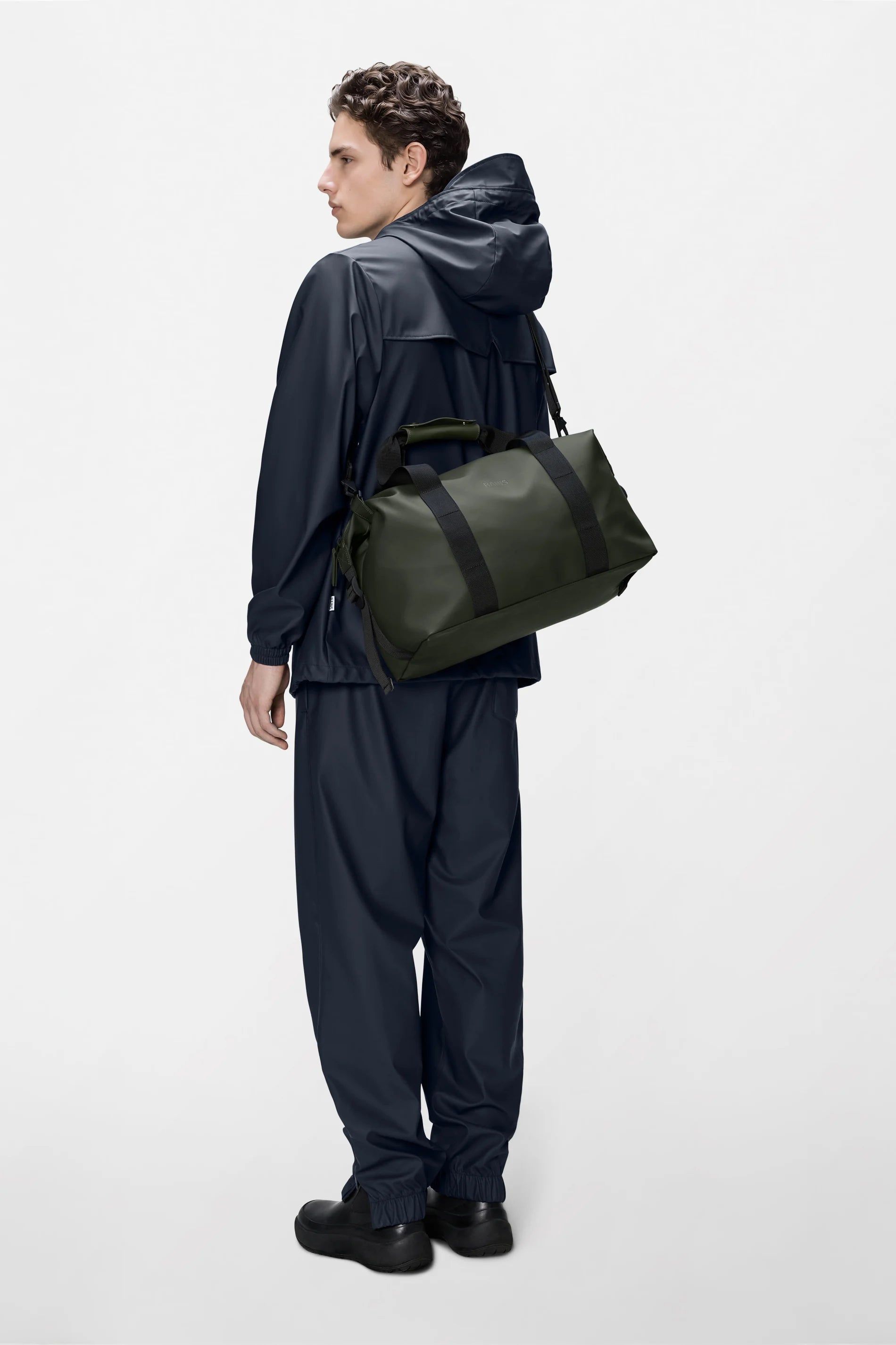 A person in a navy hooded jacket and matching pants stands facing away, carrying the Rains Hilo Weekend Bag Small in Green over one shoulder, against a plain light background.