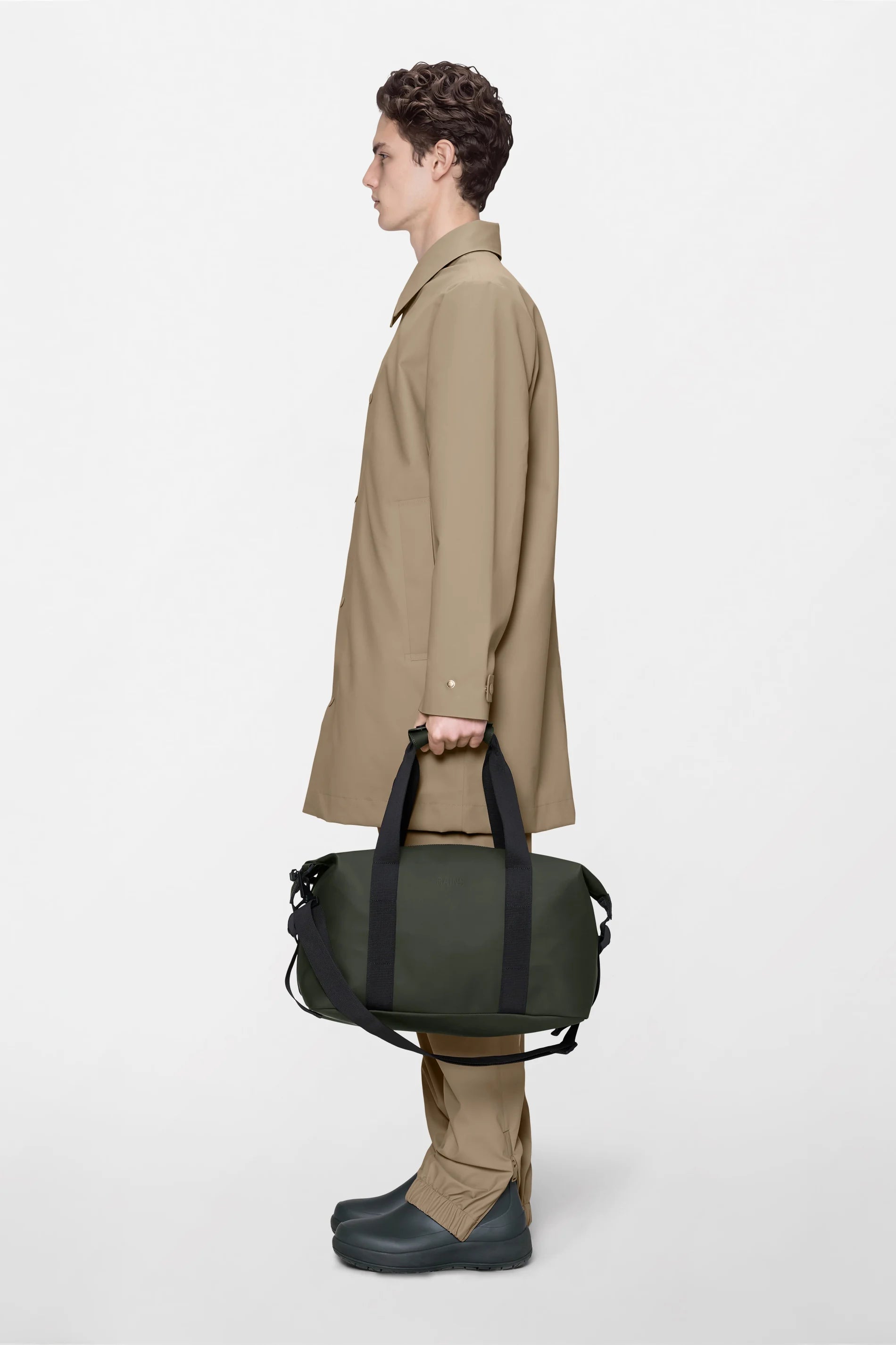 A person in a tan coat and pants stands in profile against a white background, holding the Rains Hilo Weekend Bag Small in green—a stylish, waterproof overnight bag by Rains with black straps, perfect for travel. They wear dark slip-on shoes.