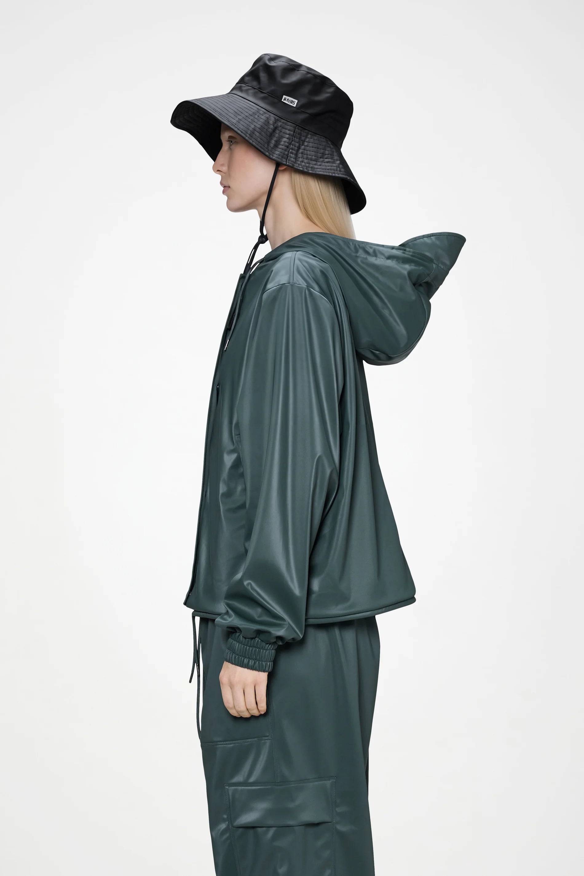 Wearing a dark green rain jacket and matching pants, a person stands in profile against a plain light background, styled with the Rains Boonie Hat in Black by Rains, featuring a chin strap.