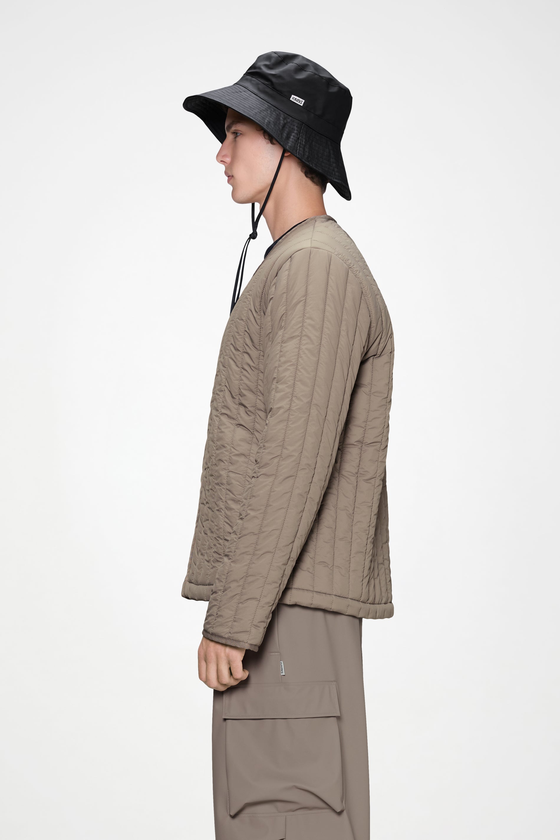 A person in a quilted beige jacket, matching cargo pants, and a Rains Boonie Hat in Black by Rains stands in profile against a plain white background.