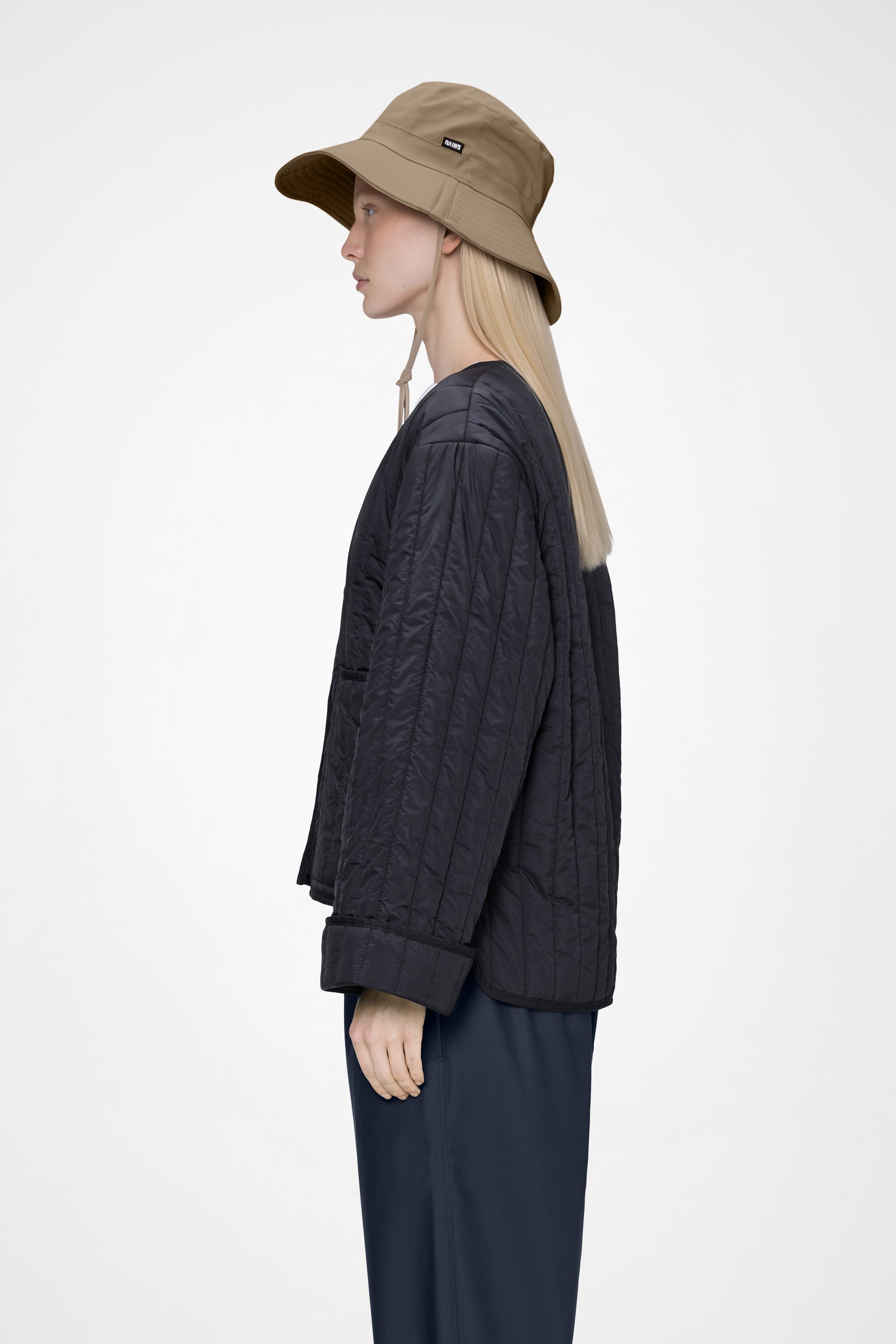 A person with long blonde hair wearing the Rains Boonie Hat in Beige by Rains, along with a dark quilted jacket and dark pants, stands in profile against a plain white background.