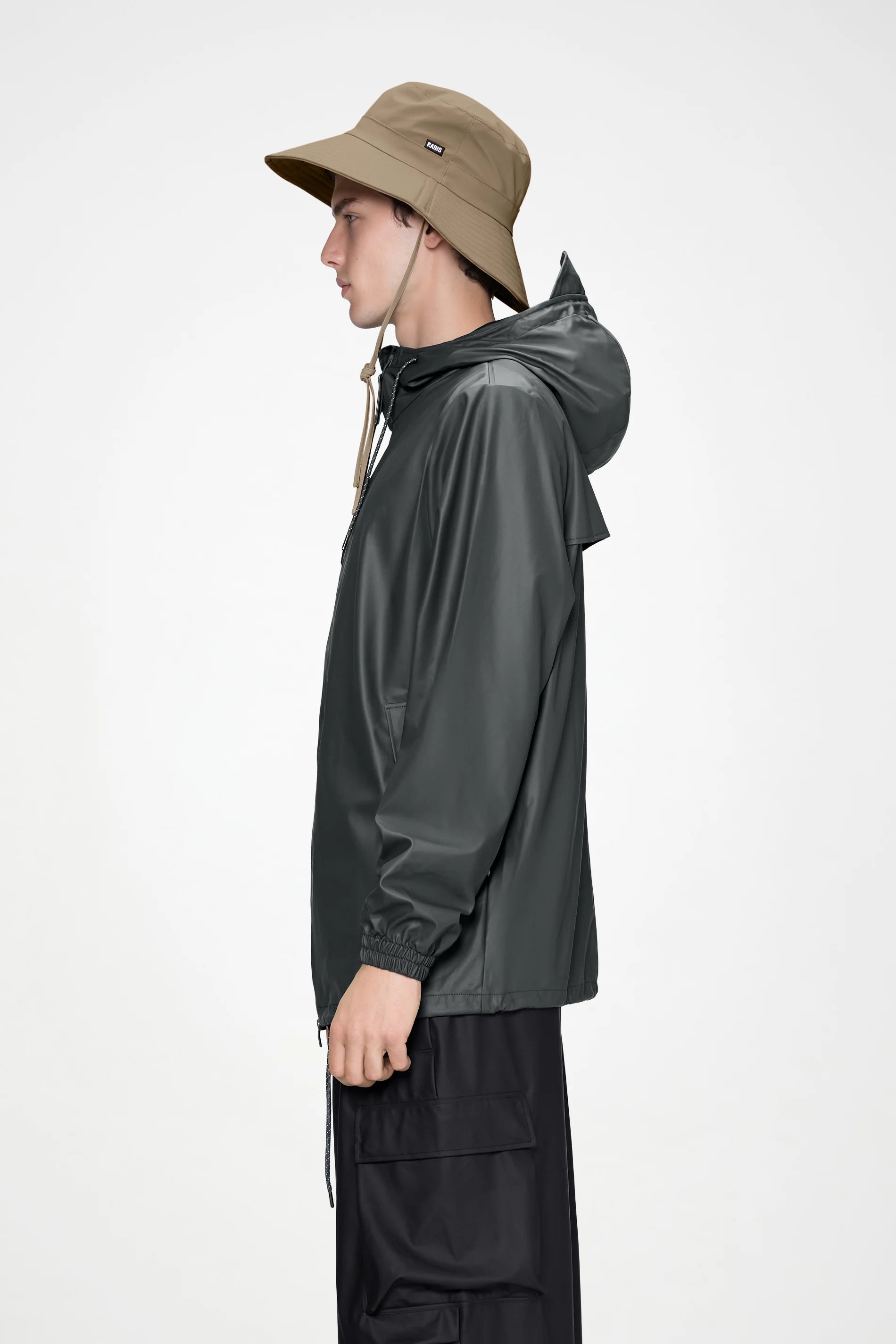 A person in a dark rain jacket made from durable PU fabric and the Rains Boonie Hat in Beige by Rains stands in profile against a plain light background.
