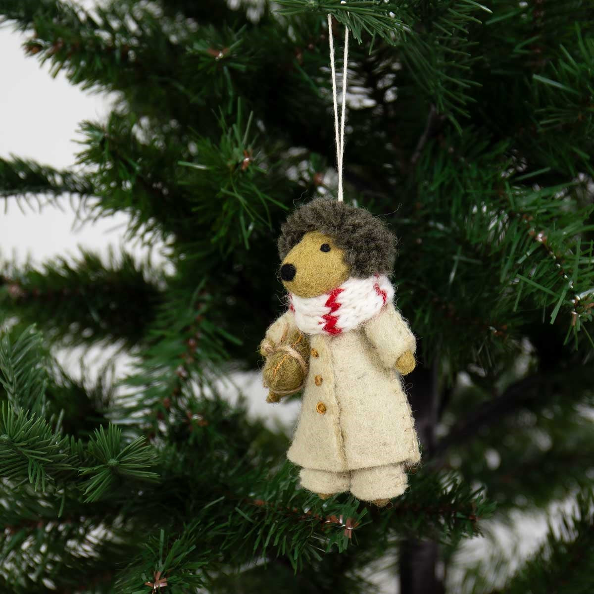 A Hedgehog in Beige Coat Christmas Ornament, Handmade in Wool, is a handcrafted felted wool hedgehog wearing a beige coat and red and white scarf, hanging from a Christmas tree branch.