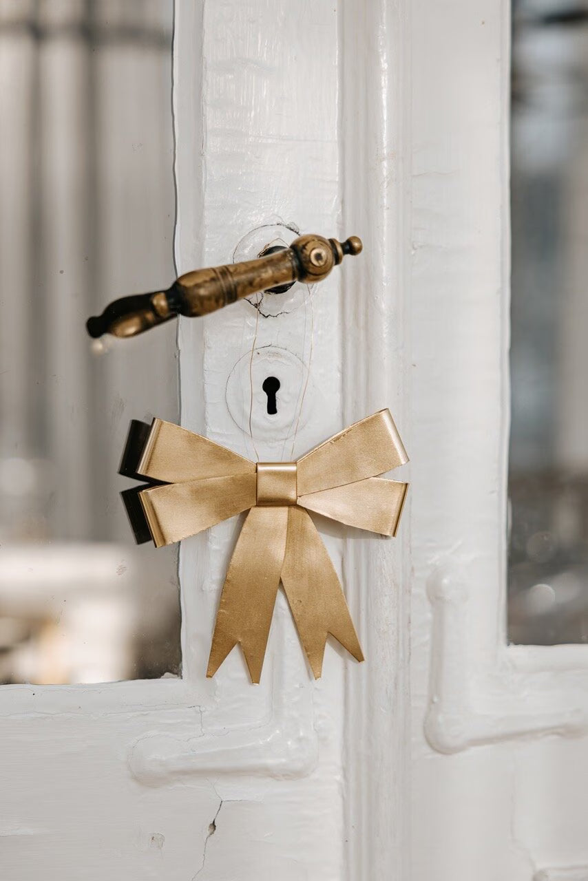 A Ribbon Hanging Ornament in brass from Strömshaga is tied around the keyhole of a white wooden door with a brass handle.