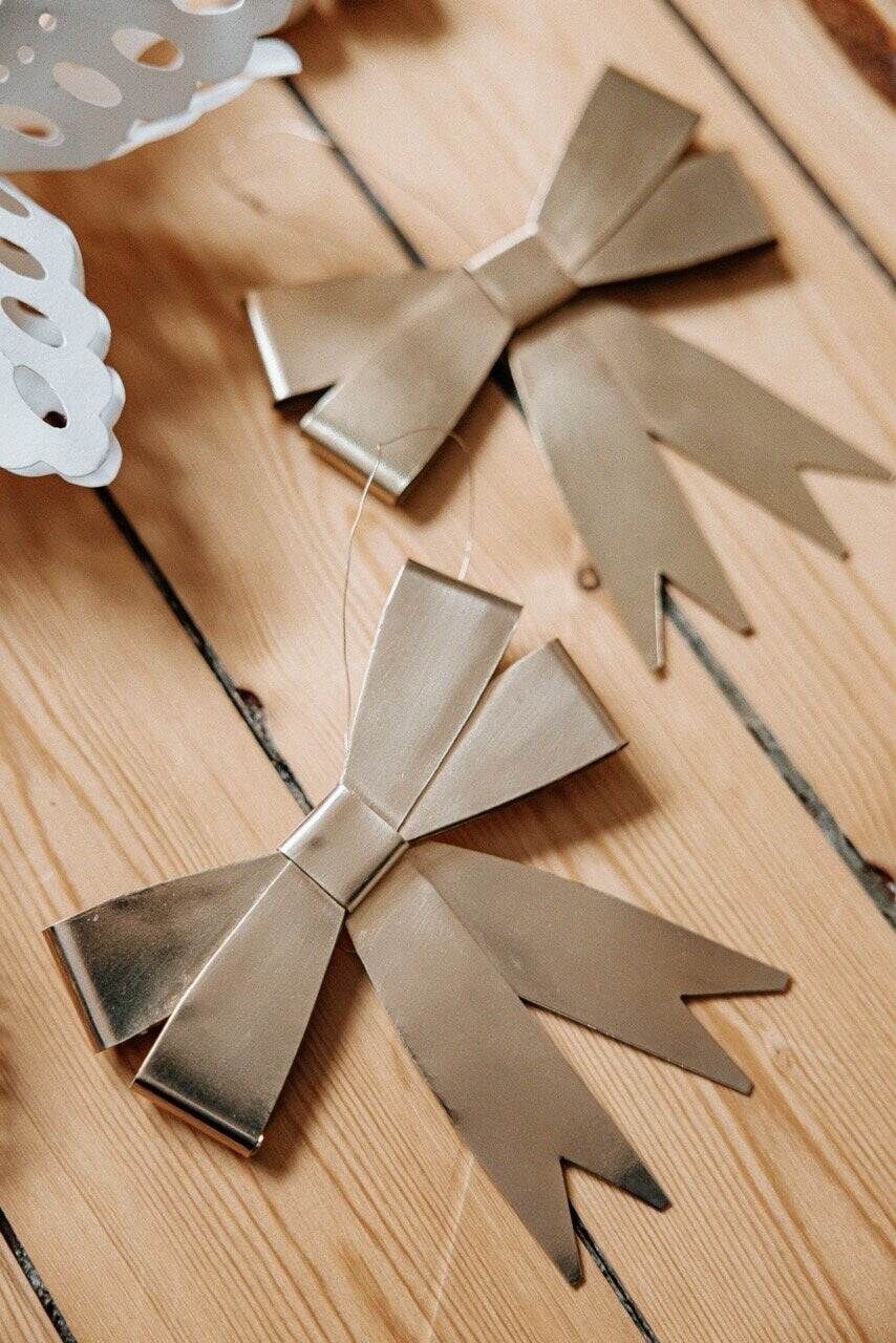 Two Ribbon Hanging Ornaments in brass from Strömshaga, featuring long, pointed tails, are arranged on a light wooden surface. In the corner, a partially visible white lacy paper decoration lends an elegant touch reminiscent of festive trimmings often seen on a Christmas tree.