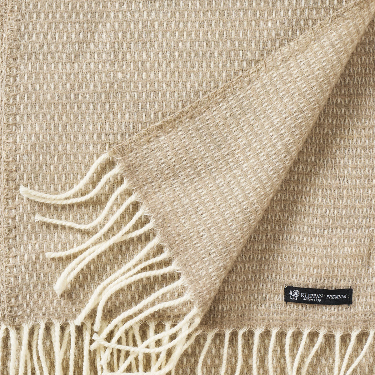 A taupe Hugo Scarf from Klippan Yllefabrik, woven from recycled and new merino wool, features white fringed edges and is shown partially folded to display both sides. A small black label with white text is sewn onto one corner, highlighting its blend of sustainable luxury.