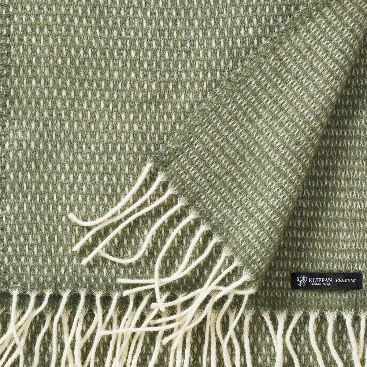 A close-up of the Hugo Scarf Recycled Merino & New Merino in Dusty Green by Klippan Yllefabrik, featuring green and white woven fabric with fringed edges. One corner is folded over to show the reverse side and a black label with “KLIPPAN” in white text.