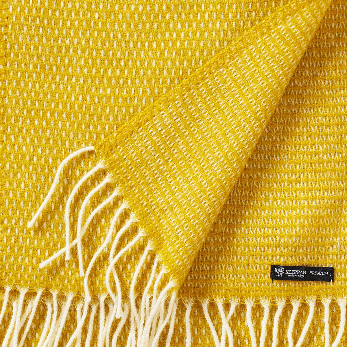 A close-up of a saffron yellow Hugo Scarf by Klippan Yllefabrik, crafted from recycled and new merino wool, features white fringe and a prominent fabric label. One corner is folded over to reveal the reverse side, beautifully showcasing its expert craftsmanship.