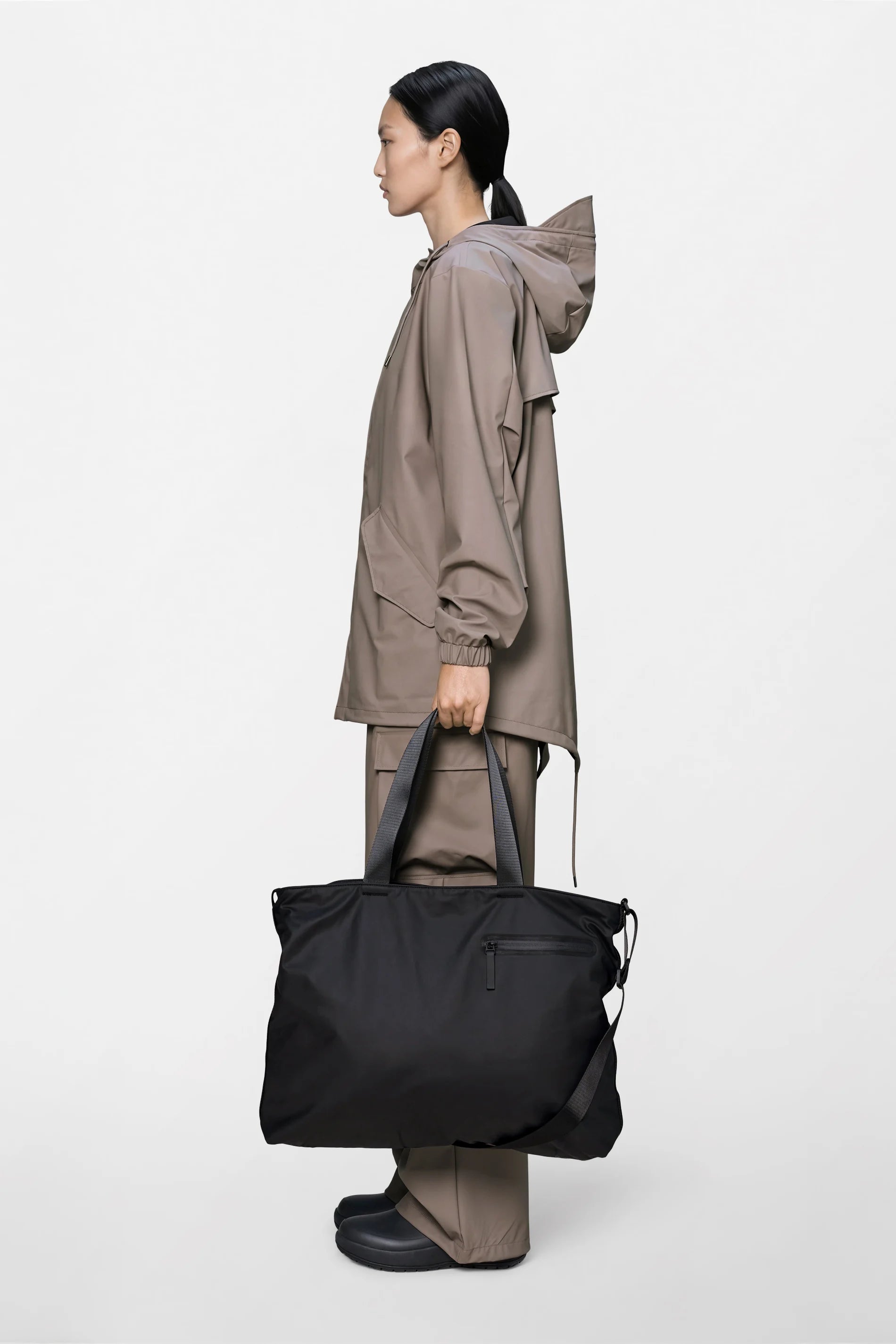 A person in a taupe rain jacket and pants stands in profile, holding the Rains Dash Messenger Tote Bag in Black by Rains. The background is plain and white.