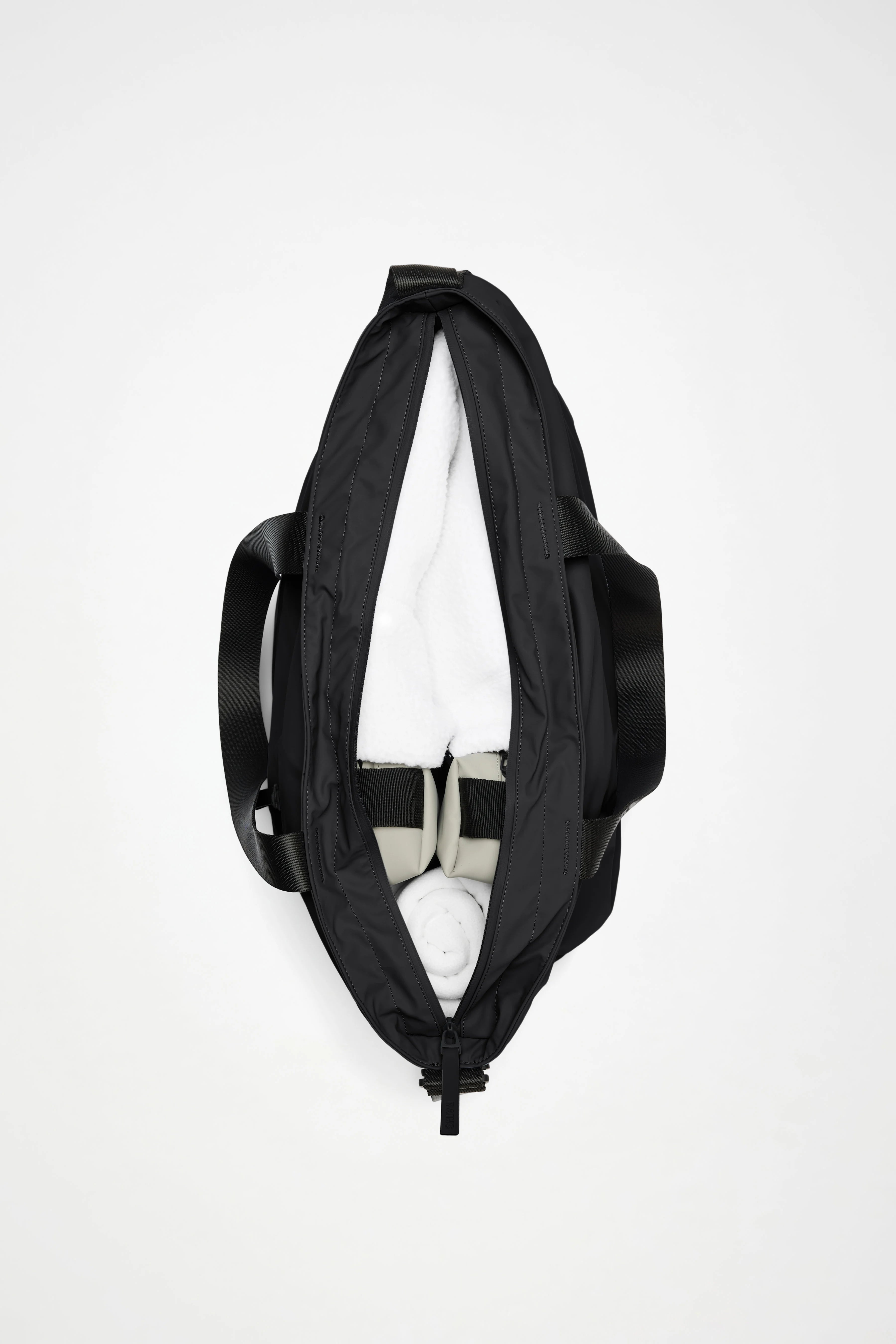 Top view of an open Rains Dash Messenger Tote Bag in Black by Rains containing a pair of white sneakers and a rolled white towel, with black handles visible on both sides.