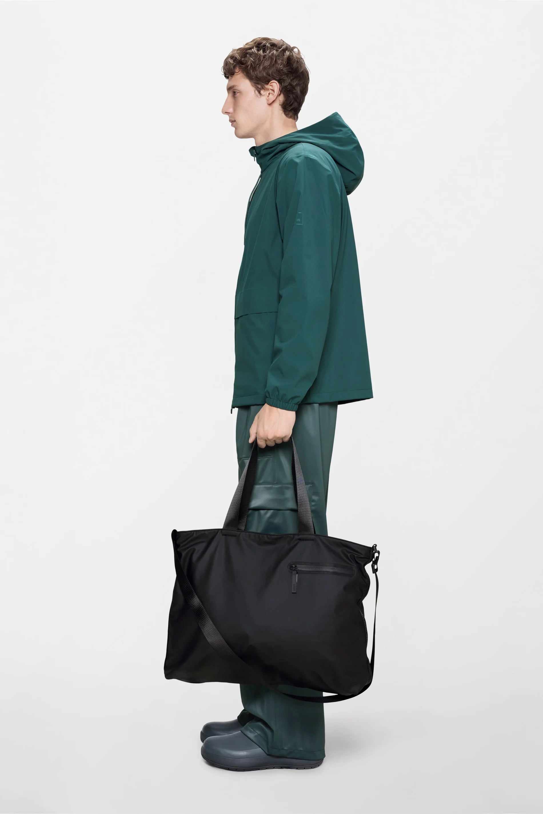 A person wearing a green hooded jacket, green pants, and gray shoes stands in profile against a plain white background, holding the Rains Dash Messenger Tote Bag in Black by Rains. The large black tote features both handles and a shoulder strap.