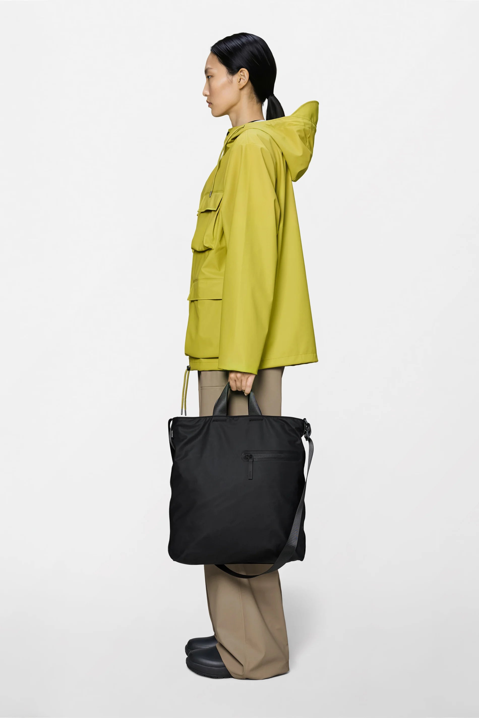 A person in a yellow hooded jacket, beige pants, and black shoes stands in profile holding the Rains Dash Tote Bag in Black by Rains, featuring an adjustable shoulder strap, against a plain white background.