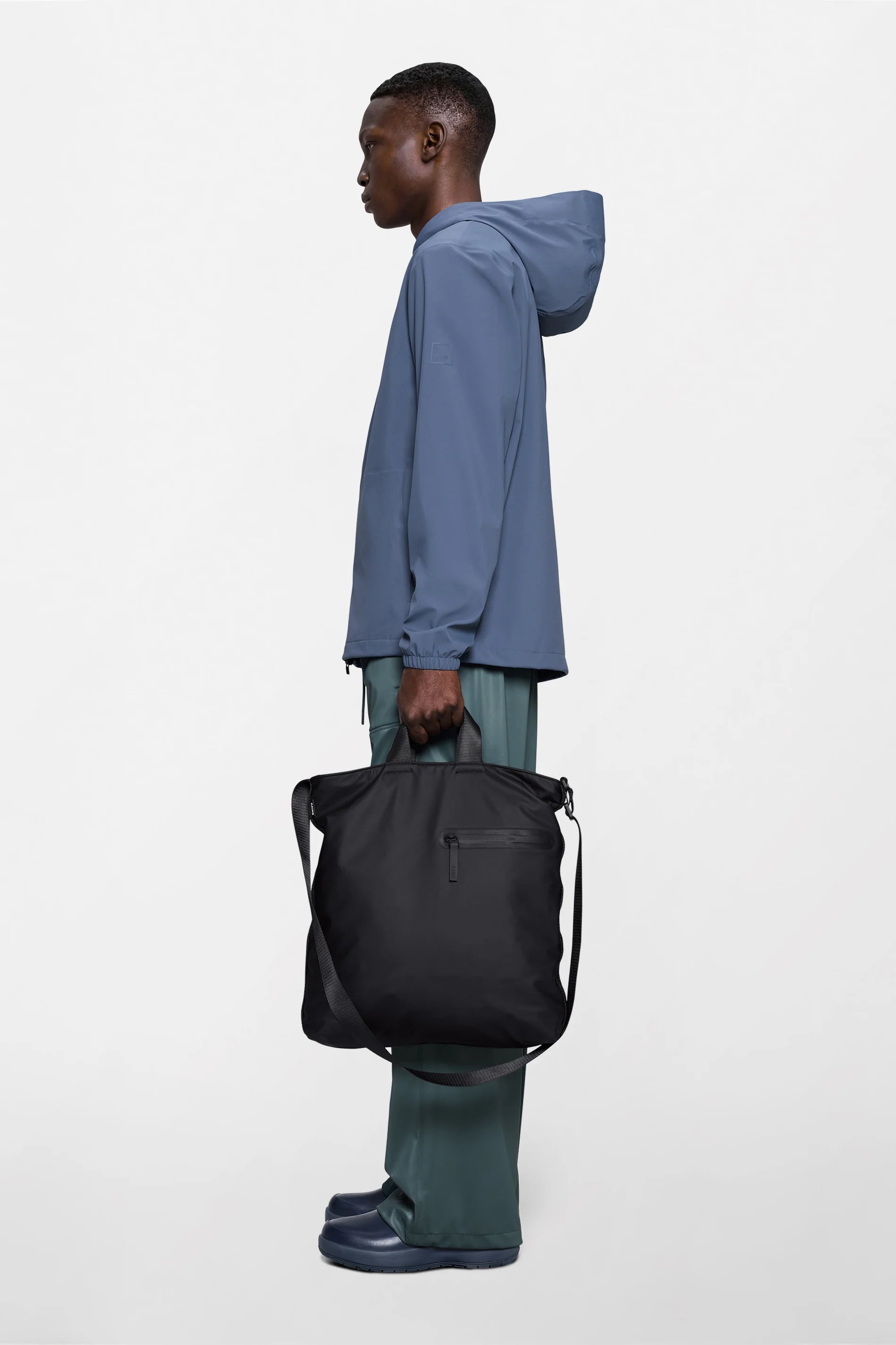 A person in a blue hooded jacket, green pants, and matching blue shoes stands in profile, holding the Rains Dash Tote Bag in Black with an adjustable shoulder strap against a plain white background.