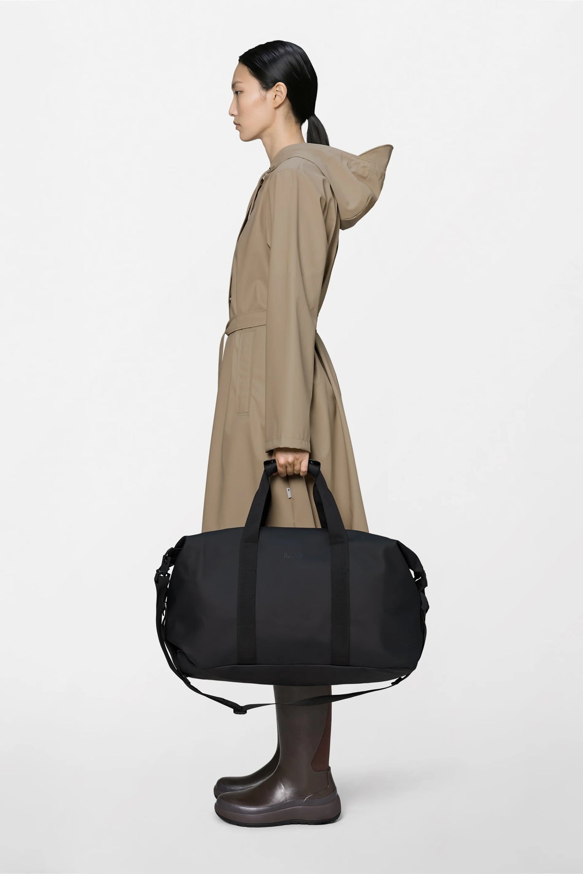 A person in a tan hooded raincoat and dark boots stands in profile, holding the Rains Hilo Weekend Bag in Black with both hands against a plain white background.