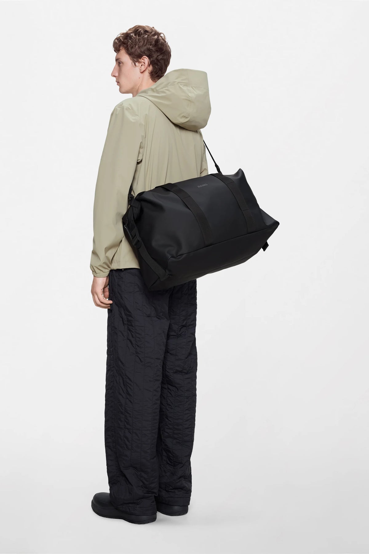 A person wearing a light beige hooded jacket and black pants stands sideways, carrying the Rains Hilo Weekend Bag in Black over their shoulder, against a plain white background.