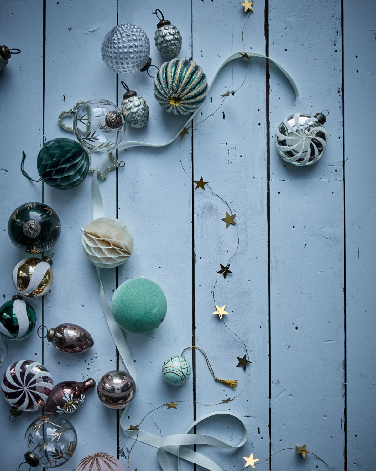 House Doctor's Garland Stars in Brass (2.5 m) is displayed on a light blue wooden surface with assorted Christmas ornaments and a ribbon woven through the festive holiday decor.