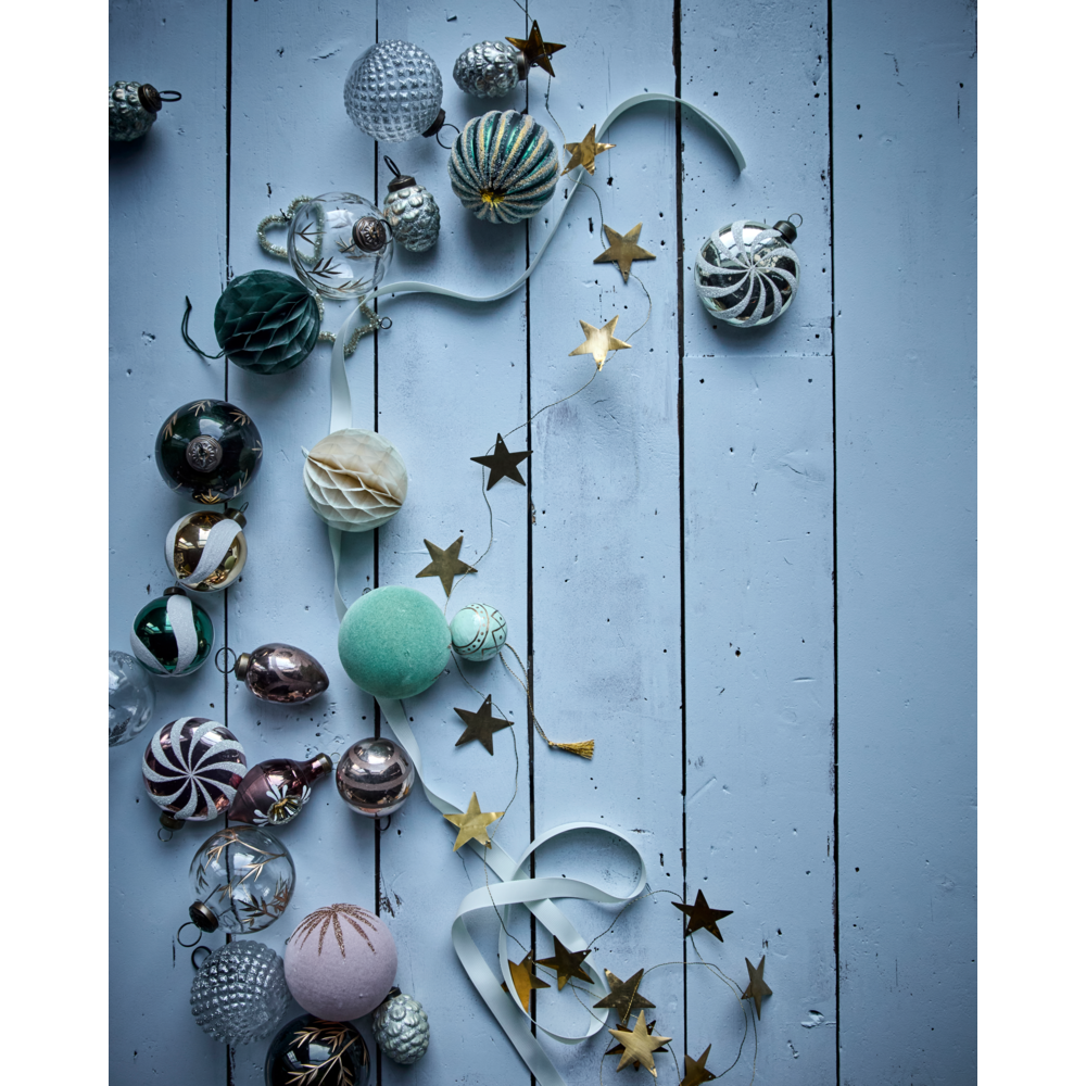 A 1.5 m Garland Stars in Antique Brass by House Doctor draped alongside assorted glass and paper Christmas ornaments on a blue wooden surface.