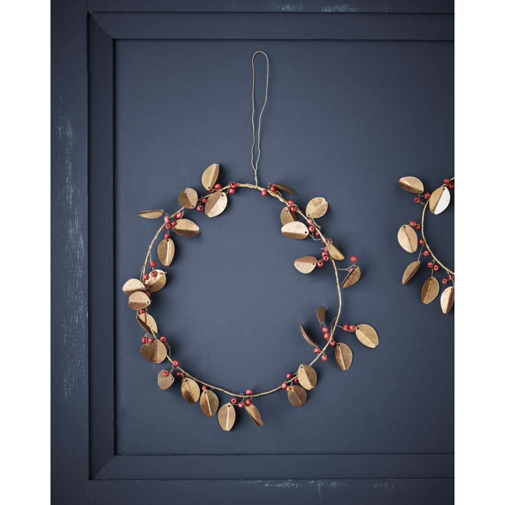 A decorative House Doctor Christmas Wreath Leafi Auric in Gold, 20 CM, made of golden brass metal leaves and small red berries, hangs as festive decor against a dark blue paneled wall.