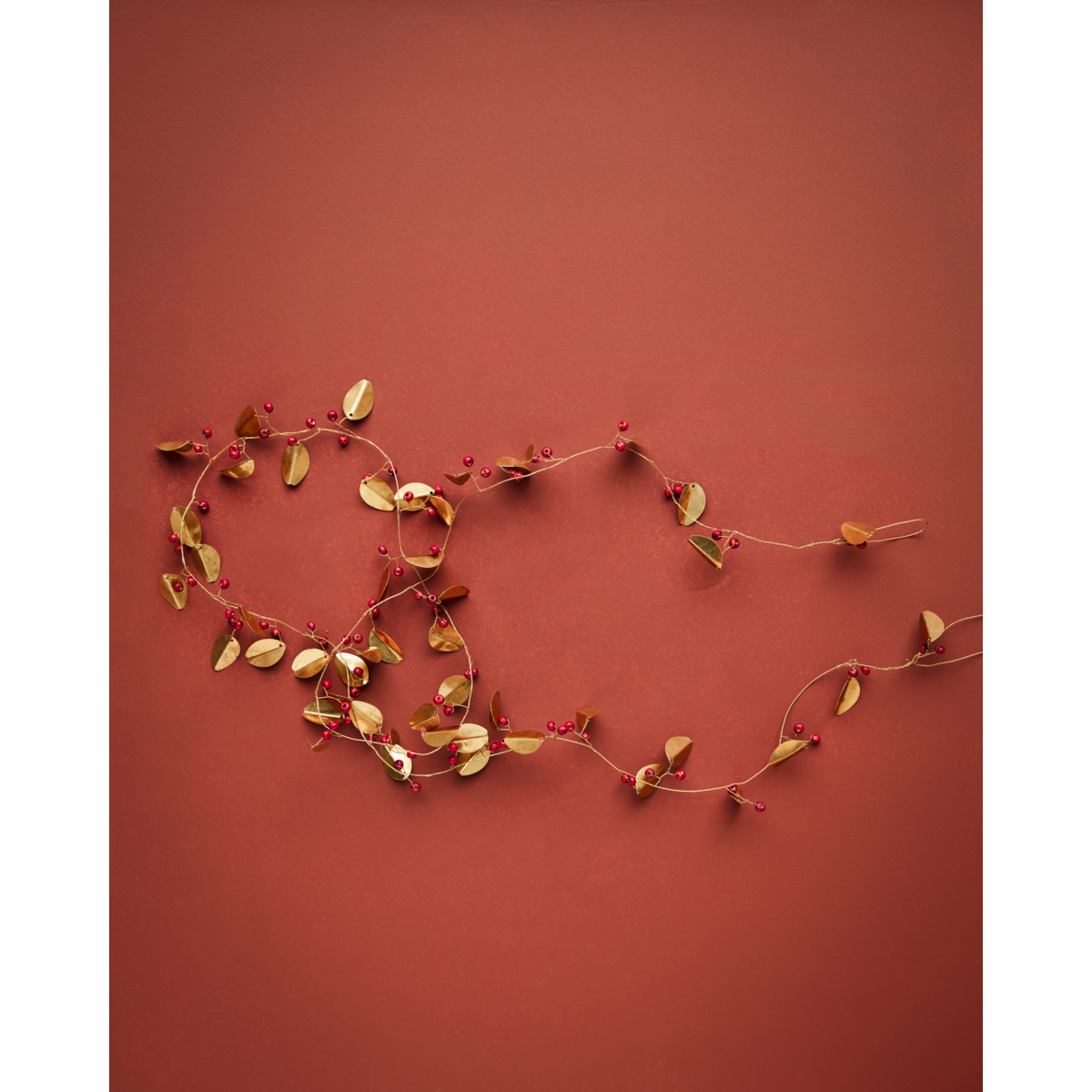 The Garland Leafi in Brass by House Doctor, measuring 183 cm, features handmade gold-toned leaves and small red berries arranged in a loose spiral on a red background—an ideal accent for festive Christmas décor.