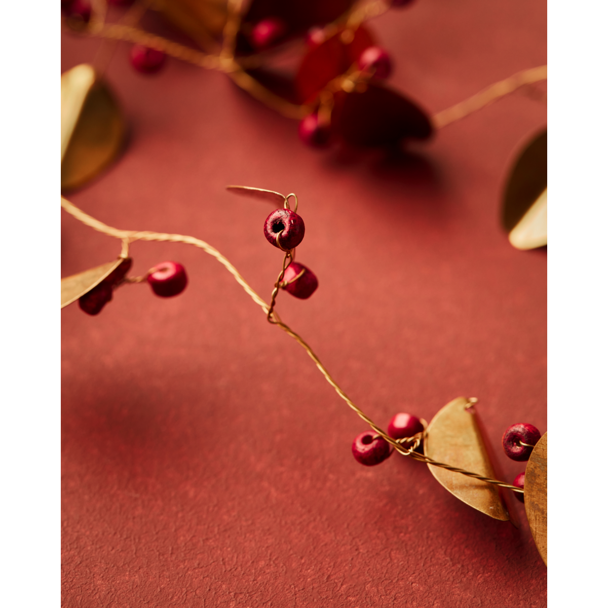 A close-up of House Doctor’s Garland Leafi in Brass (183 cm) showcases shiny red berries and golden leaves on a textured, dark red surface, with a softly blurred background highlighting this festive Christmas décor.
