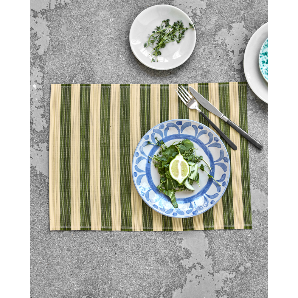 A House Doctor "Rayas" Green & Nature Stripe placemat from a set of 4 is shown on a gray surface, topped with a blue and white plate of leafy greens and lemon slices, cutlery, and two small dishes including one with herbs.