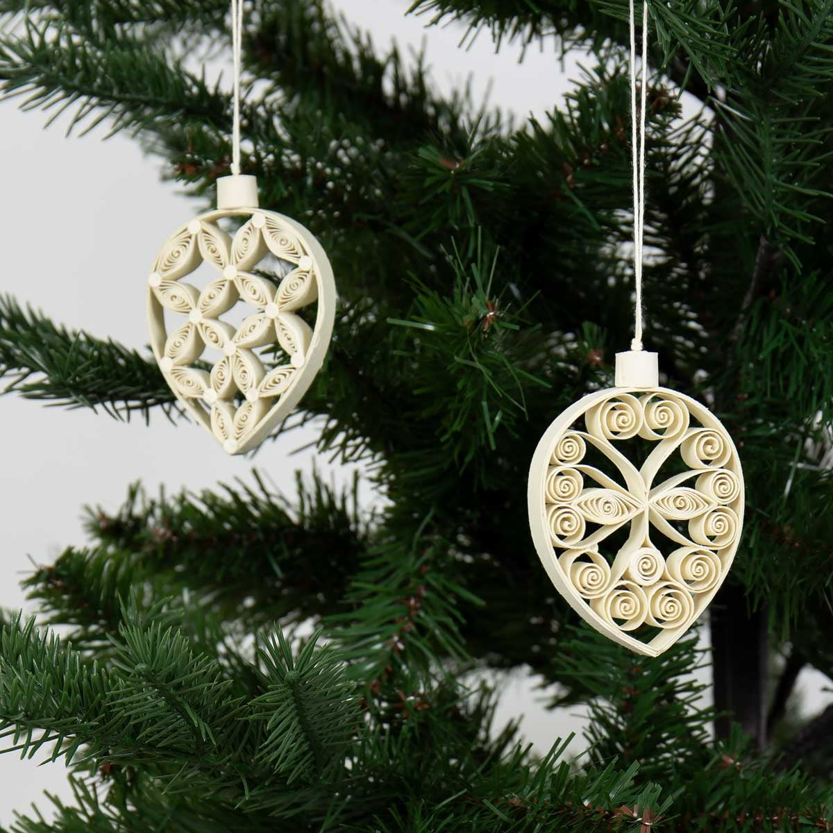 Two beige Quilled Leaf Tree Christmas Ornaments by A World of Craft hang on the green branches of an artificial Christmas tree against a light background, highlighting the delicate artistry of these handmade paper quilling decorations and their timeless charm.