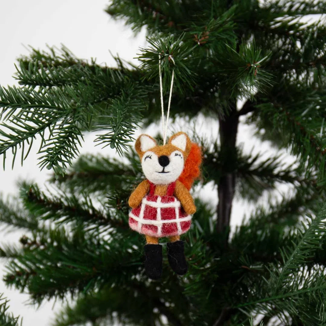 The Shy Fox Christmas Ornament, a handcrafted felt fox wearing a red plaid dress and black boots, hangs from a pine branch with woodland charm.