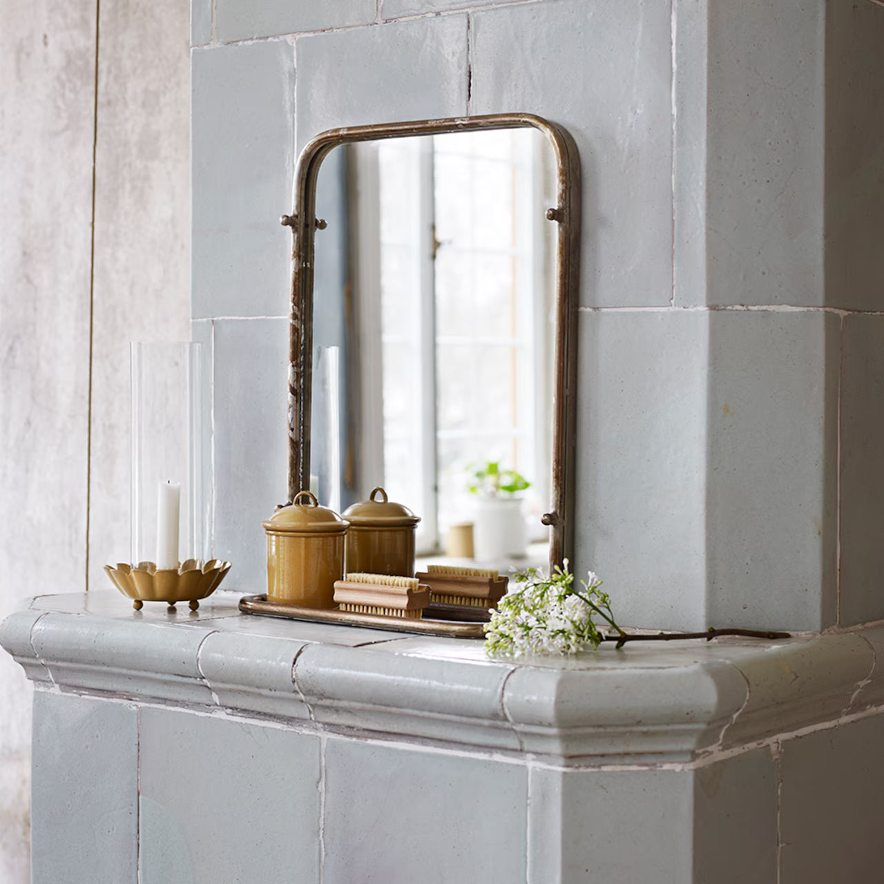 A mantel with a vintage mirror, two brown jars, the Strömshaga Nailbrush in Ash Wood, a gold candle holder with a white candle, and a small bunch of white flowers.