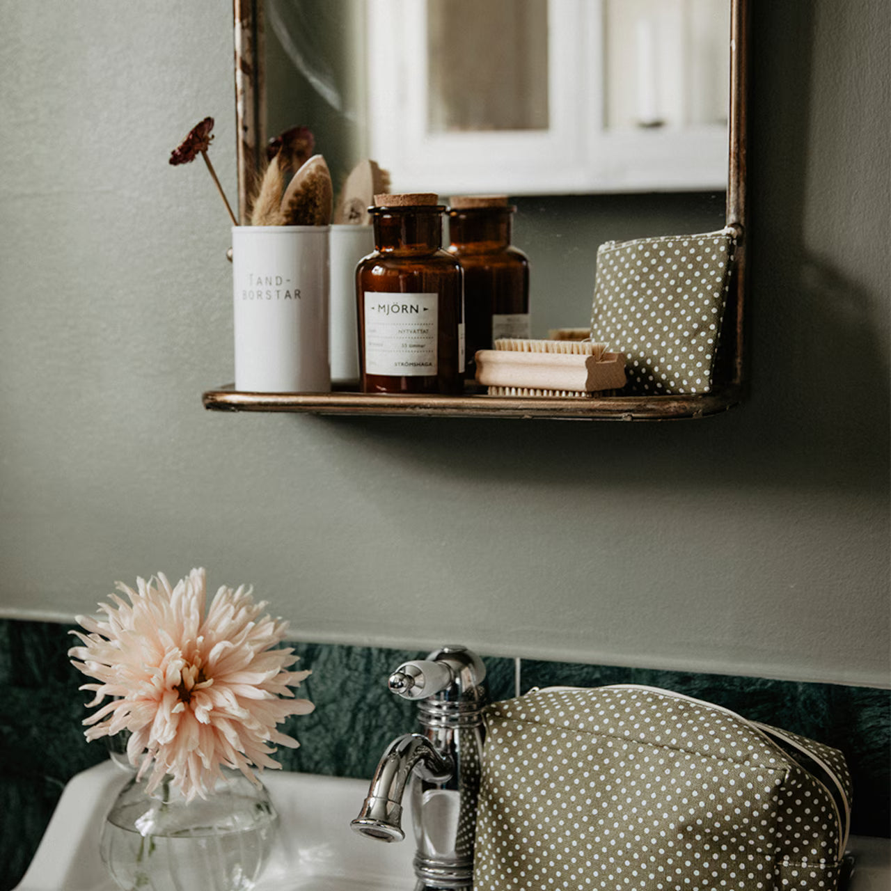 On the bathroom sink sits Strömshaga’s Nailbrush in Ash Wood, a polka dot toiletry bag, a vase with a flower, and a shelf with bottles and a toothbrush holder, all reflected in the mirror.
