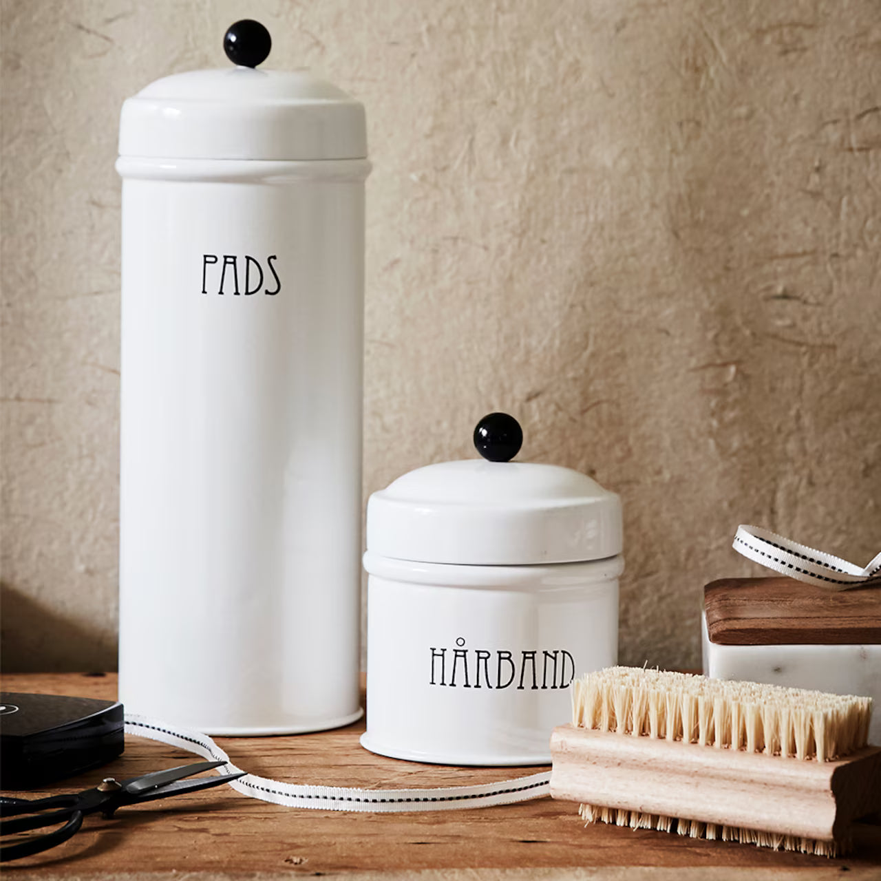 Two white ceramic containers labeled Pads and Hårband sit on a wooden surface beside the Strömshaga Nailbrush in Ash Wood, tampico fibre, a ribbon, and grooming tools, all set against a textured beige background.