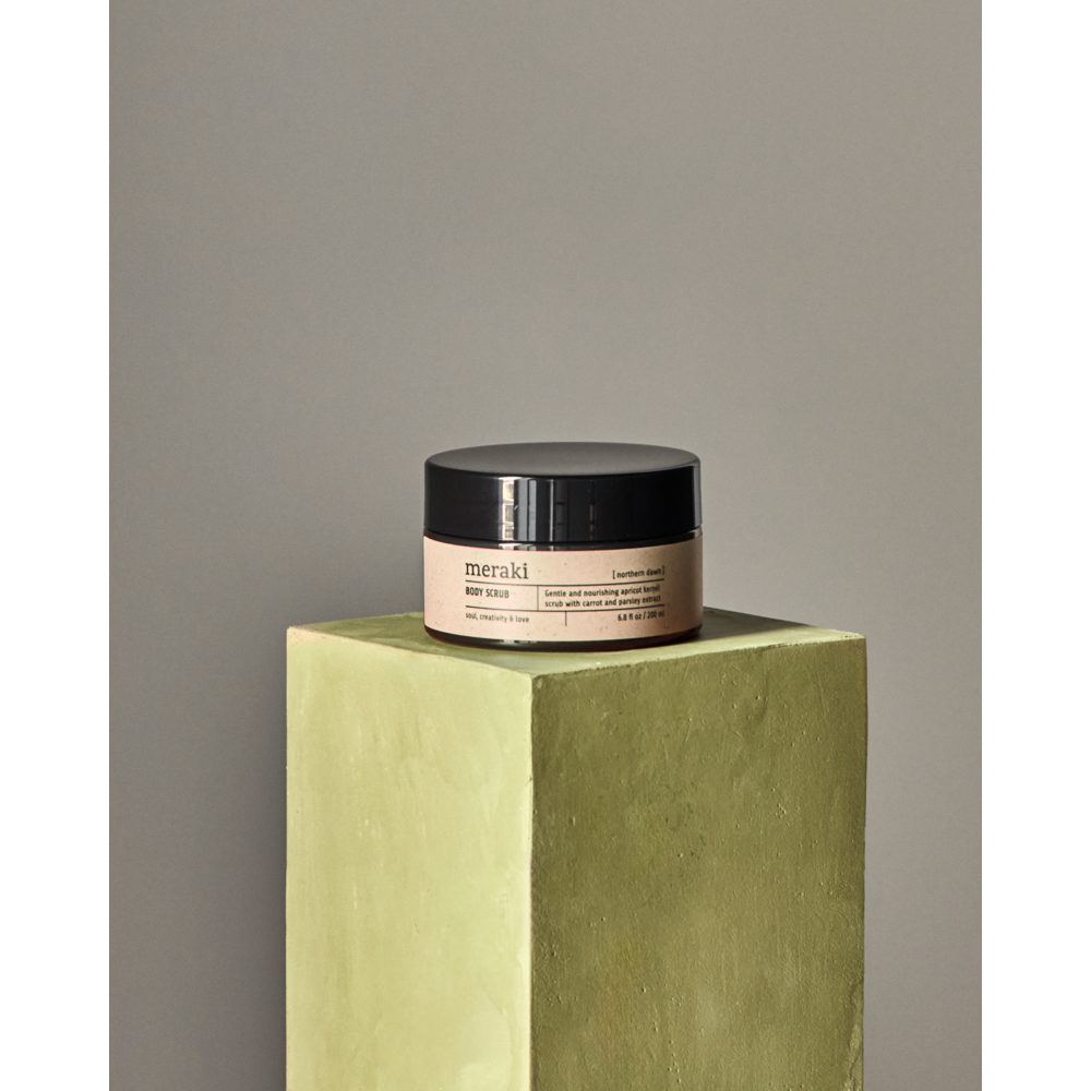 A jar of Meraki Body Scrub Organic, Northern Dawn with a black lid sits on a green rectangular pedestal against a plain, neutral background.