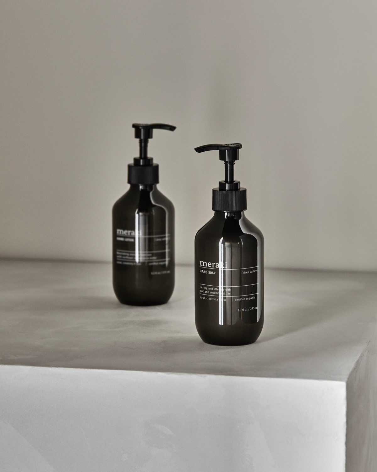 Two dark glass pump bottles from the Meraki Gift Box Deep Valley Hand Care Set—with organic hand soap and lotion—sit on a minimalist light gray surface. One bottle is in focus, highlighting this elegant gift choice.