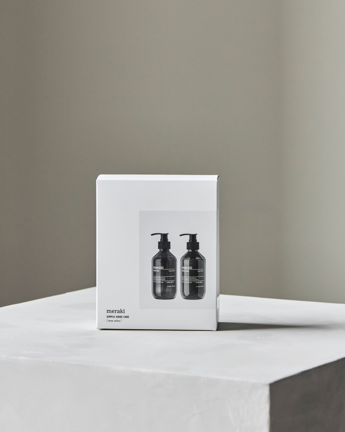 A white gift box featuring an image of two black Meraki bottles, the Gift Box Deep Valley Hand Care Set with Hand Soap & Hand Lotion, sits on a gray surface—an ideal present for fans of organic hand care.