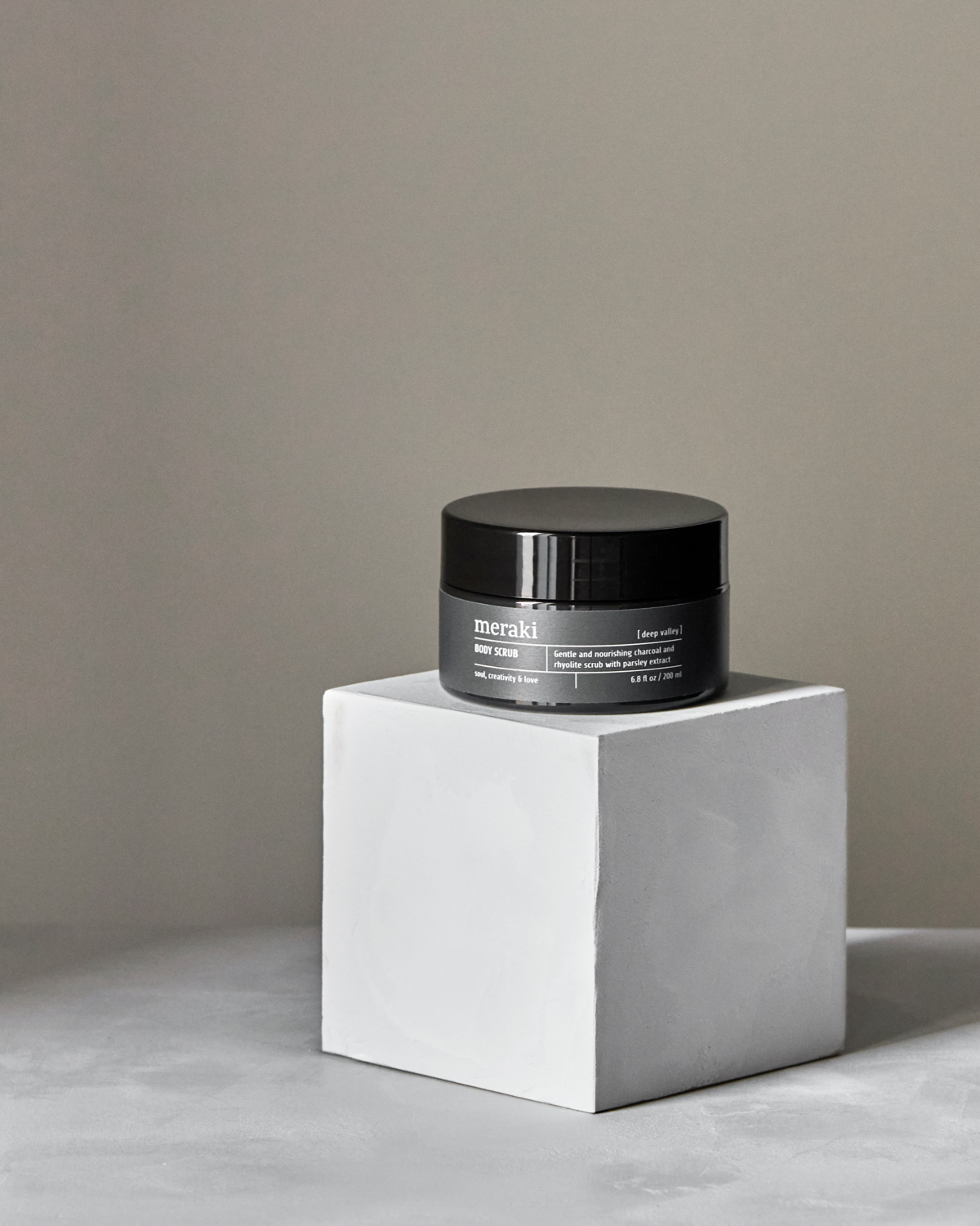 A 200 ml jar of Meraki Body Scrub, Deep Valley rests on a white cube pedestal against a neutral gray background, offering gentle exfoliation for refreshed skin.
