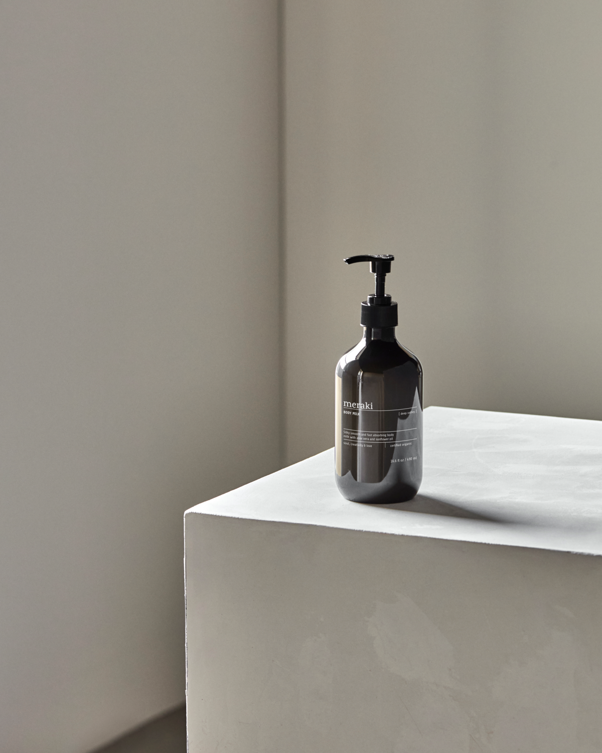 A dark pump bottle of Meraki Body Milk, Deep Valley, 490 ml rests on a light gray minimalist pedestal in a softly lit room with neutral walls.