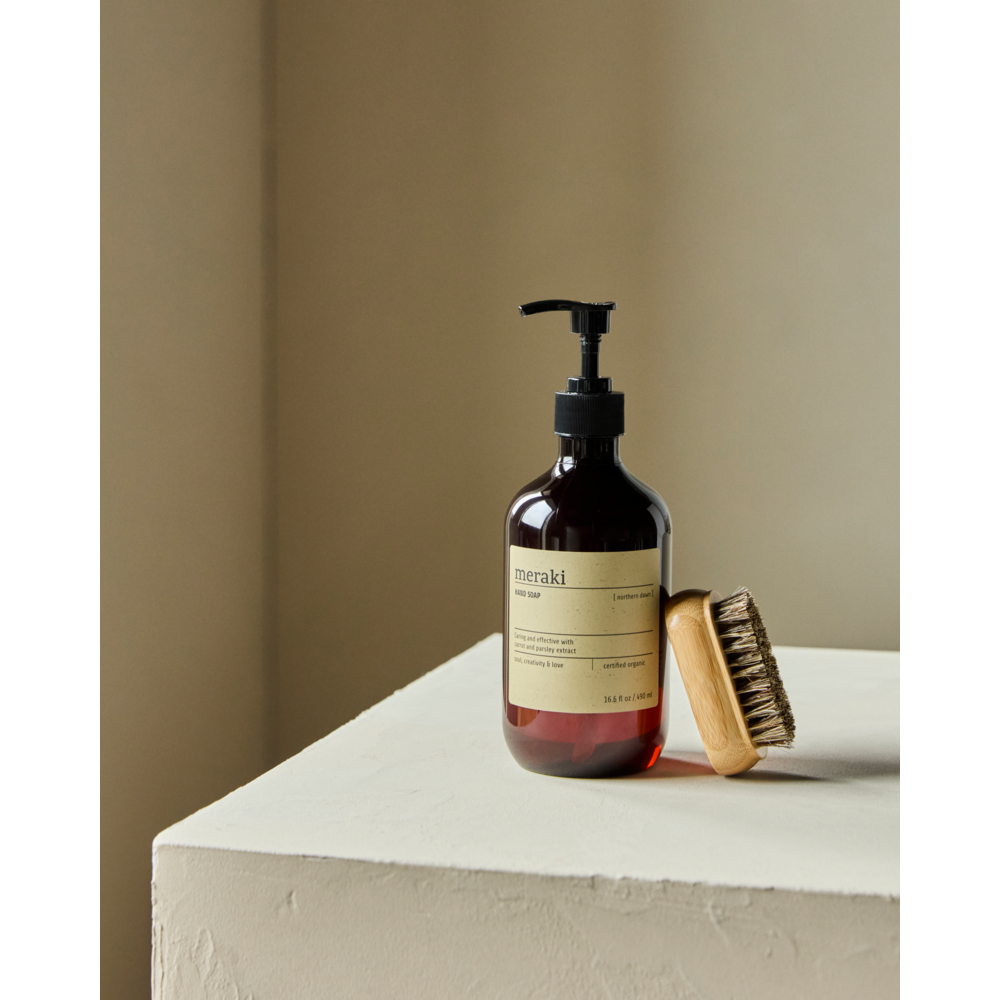 A brown Meraki Hand Soap Organic, Northern Dawn bottle and a wooden bristle brush rest on a light textured surface with a neutral beige backdrop.