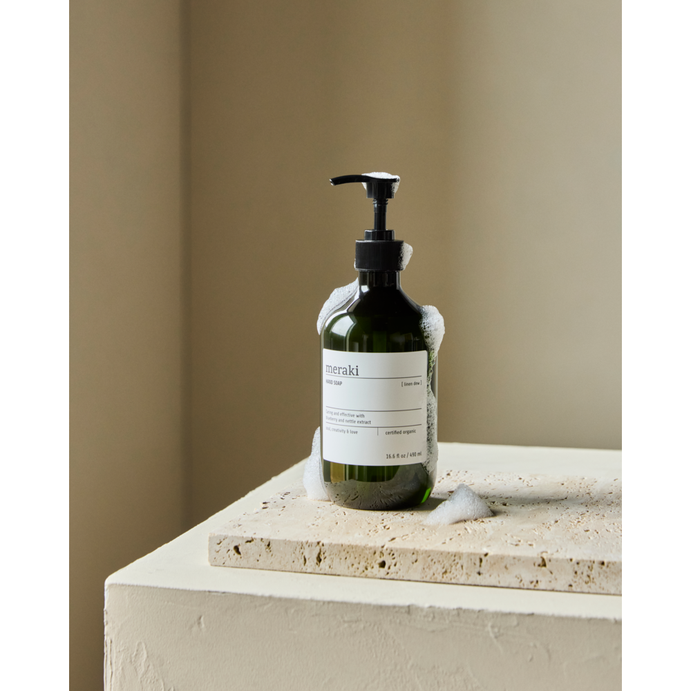 A green pump bottle of Meraki Hand Soap Organic, Linen Dew stands on a beige stone slab with soap suds around its base, set against a soft beige background. Enriched with blueberry extract and certified organic ingredients.