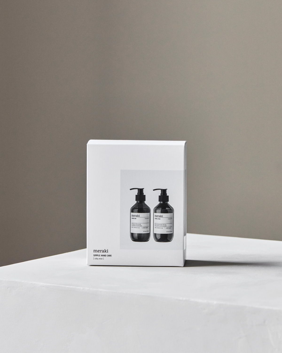 A white, minimalist Meraki gift box featuring two black pump bottles and "Gift Box Silky Mist, Hand Care Set with Hand Soap & Hand Lotion" text sits on a white surface against a beige background, suggesting premium hand care inside.