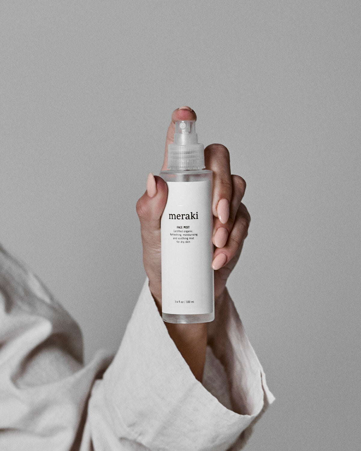 A hand with pale pink nails holds a clear Meraki Face Mist bottle, Organic with Earth Marine Water, against a neutral gray background. The person wears a light-colored, long-sleeved top—perfect for achieving a radiant complexion.