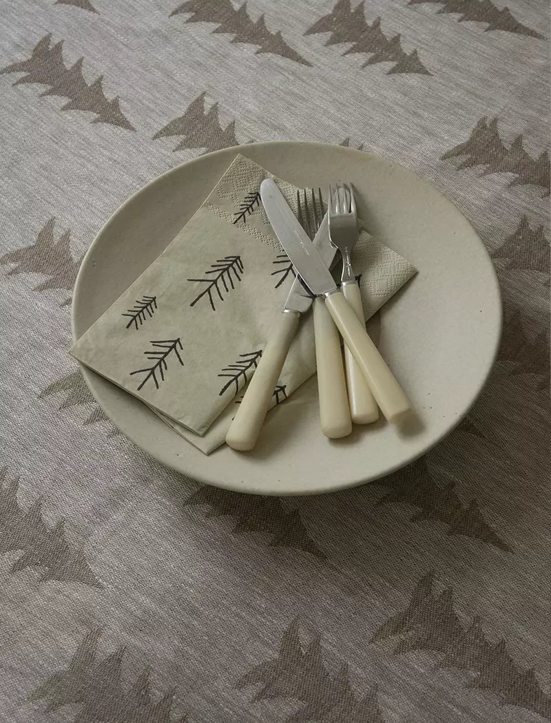 A cream-colored plate displays a Fine Little Day Pine (Tall) Paper Napkin (33x33 cm) with pine tree print, paired with cutlery featuring ivory handles on a beige patterned tablecloth—a refined touch for any table setting.