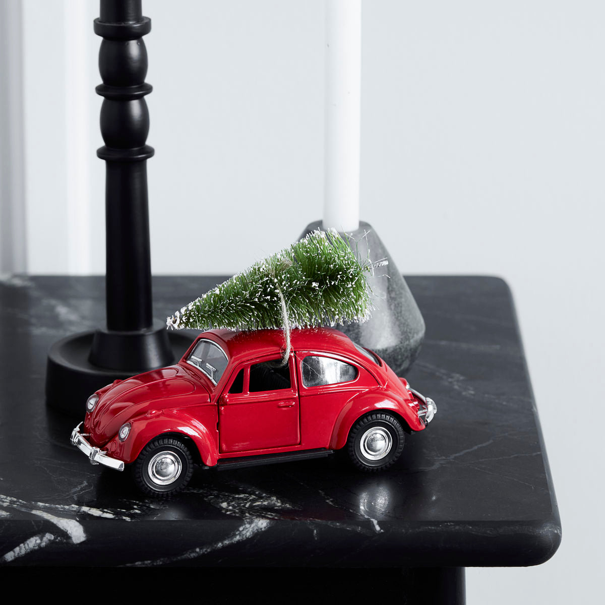 The House Doctor Ornament Xmas Car Christmas Decoration in Red, featuring a miniature tree on its roof, sits on a black marble surface beside candle holders, adding nostalgic charm to your holiday décor.
