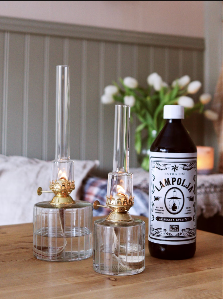 Two elegant glass kerosene lamps with brass tops sit on a wooden table beside a dark Strömshaga Lamp Oil 1 Liter bottle; in the softly blurred background is a vase of white tulips.